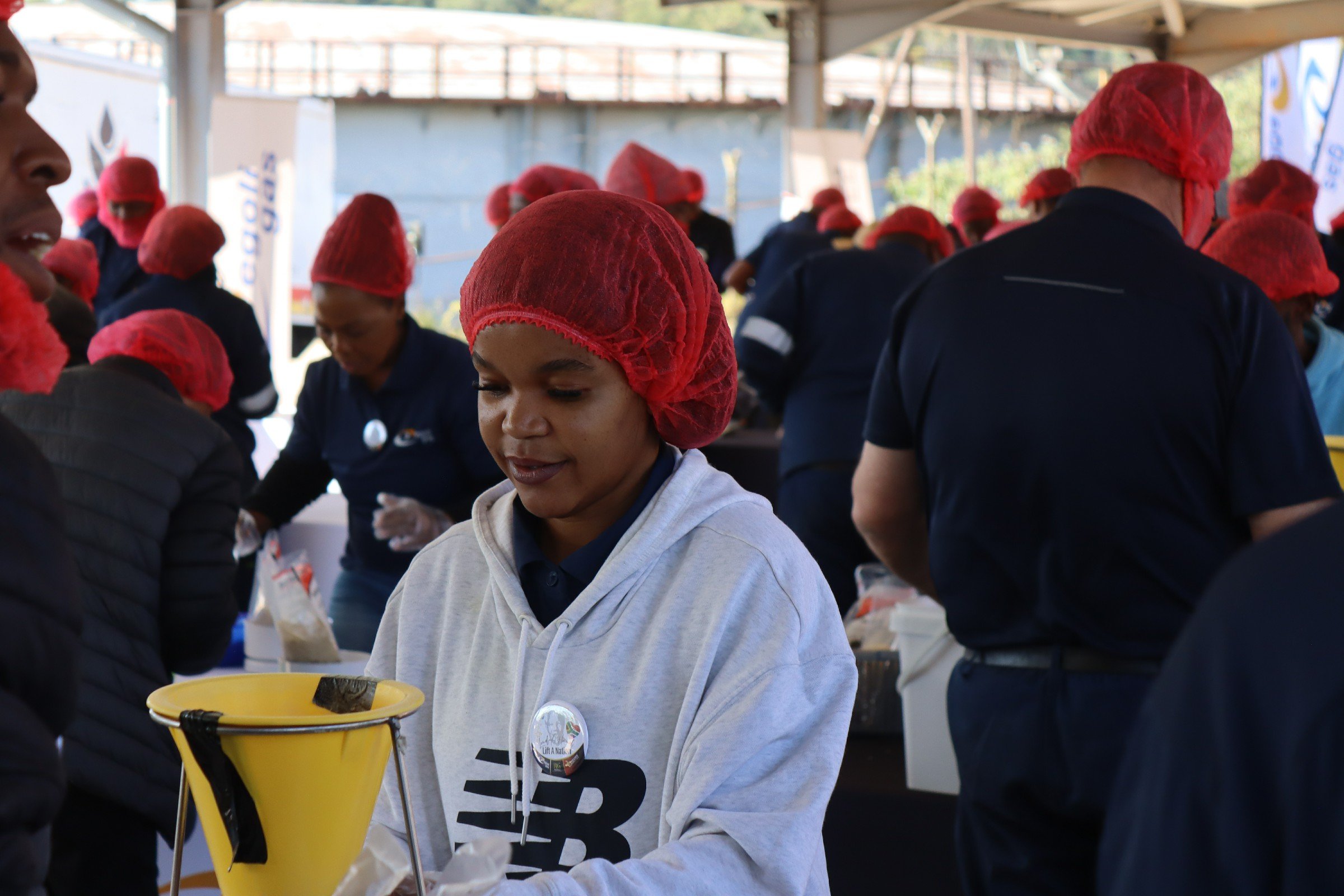A woman wearing a red hairnet and a gray hoodie working at a food distribution event indoors, with several people in the background also wearing red hairnets and safety gloves, under a covered outdoor area.
