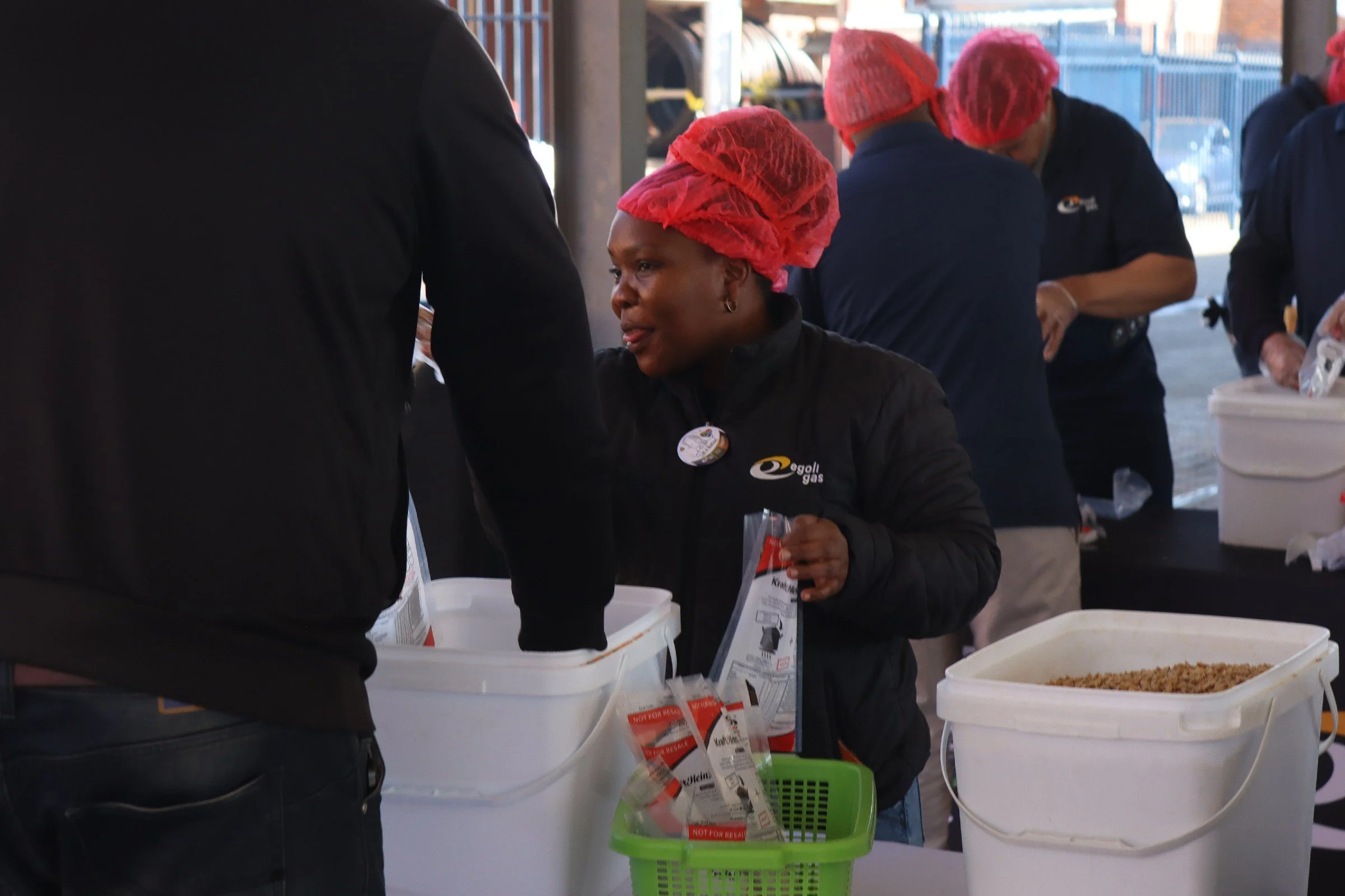 Women wearing red hairnets working at a table with food supplies and informational flyers in an outdoor setting.
