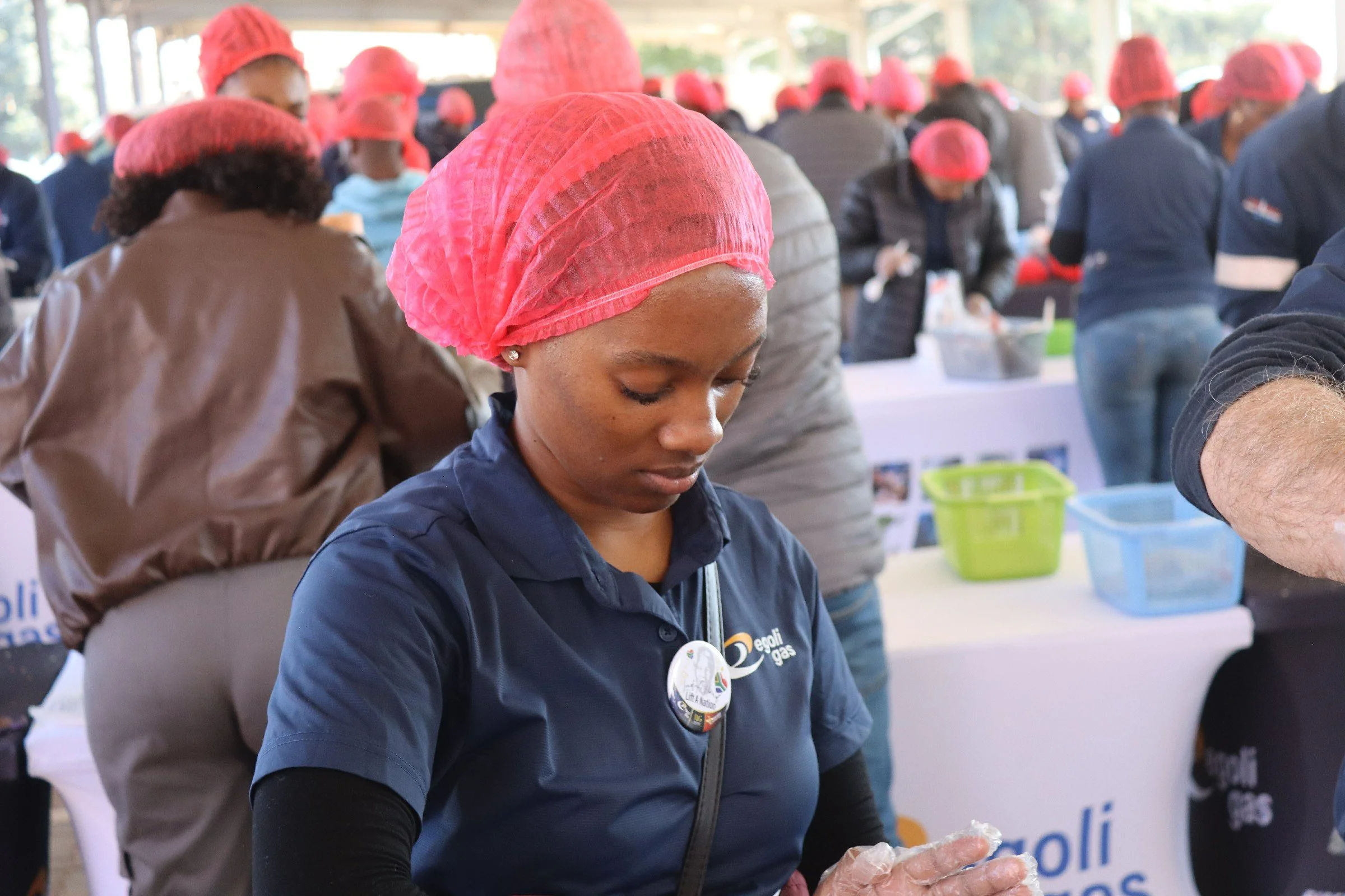 A woman wearing a pink hair cover and a navy blue shirt with a logo, working at an indoor event with a crowd in the background, many also wearing pink hair covers.