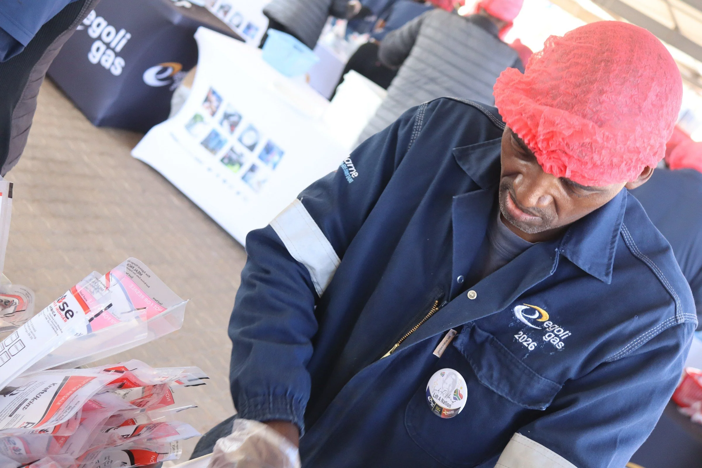 A man wearing a red hat and a dark blue jacket with a badge on it is standing at a table at an event. There are pamphlets on the table, and other people are visible in the background.