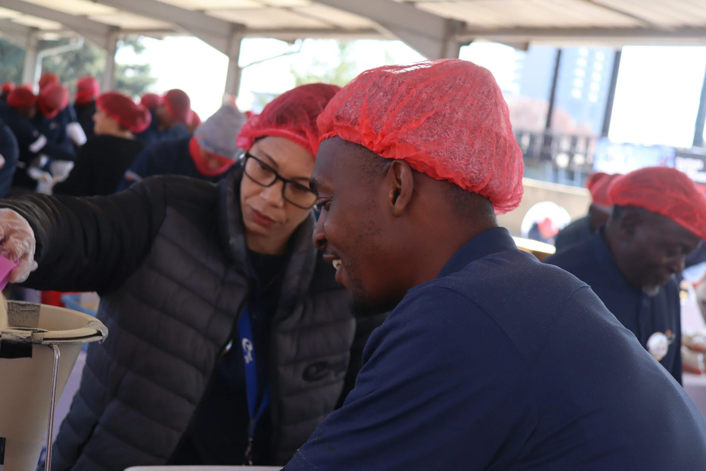 People wearing red hairnets and dark clothing working together at a table indoors with large windows showing city buildings in the background.