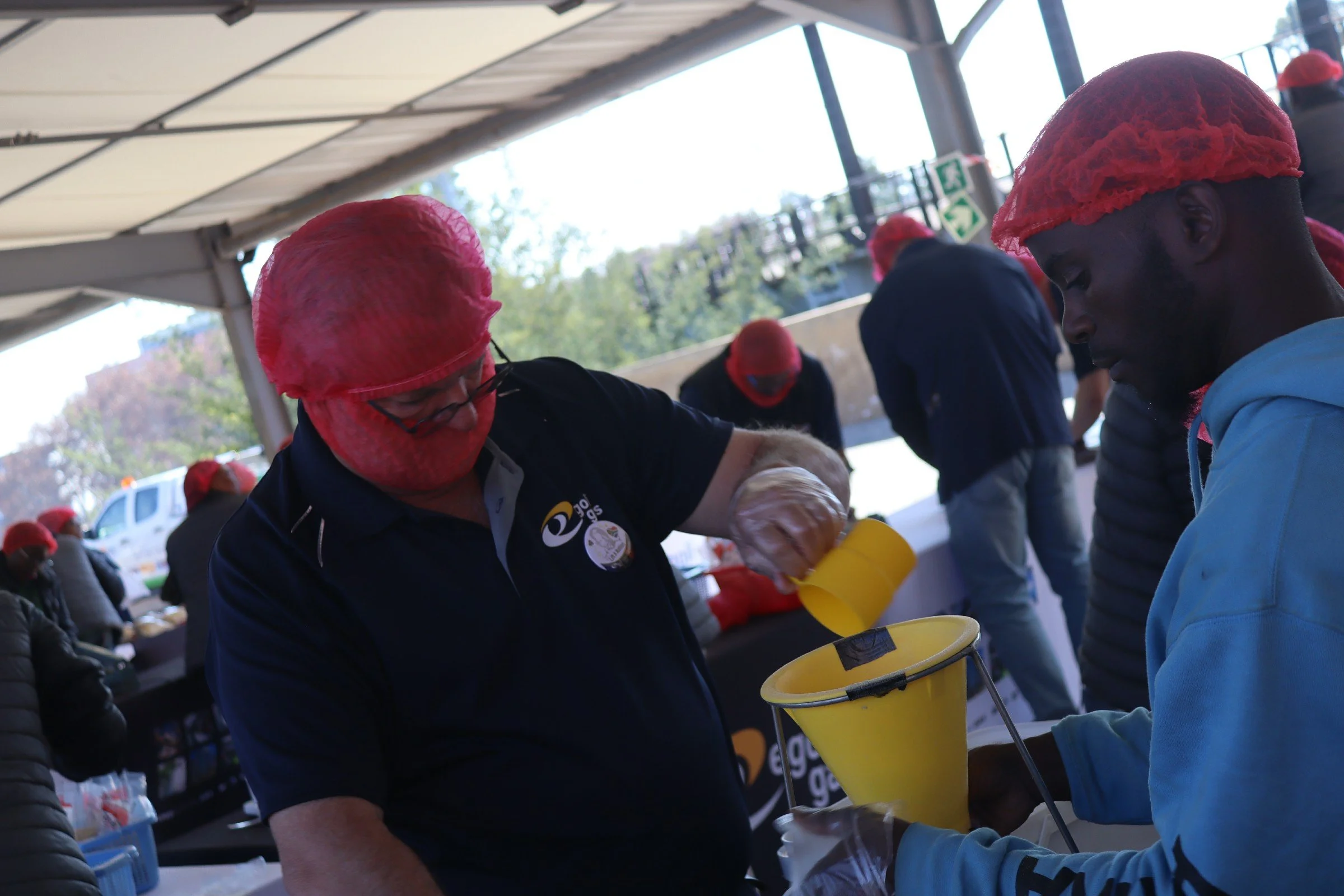 People wearing red hairnets distributing food at a community event under a covered outdoor area.