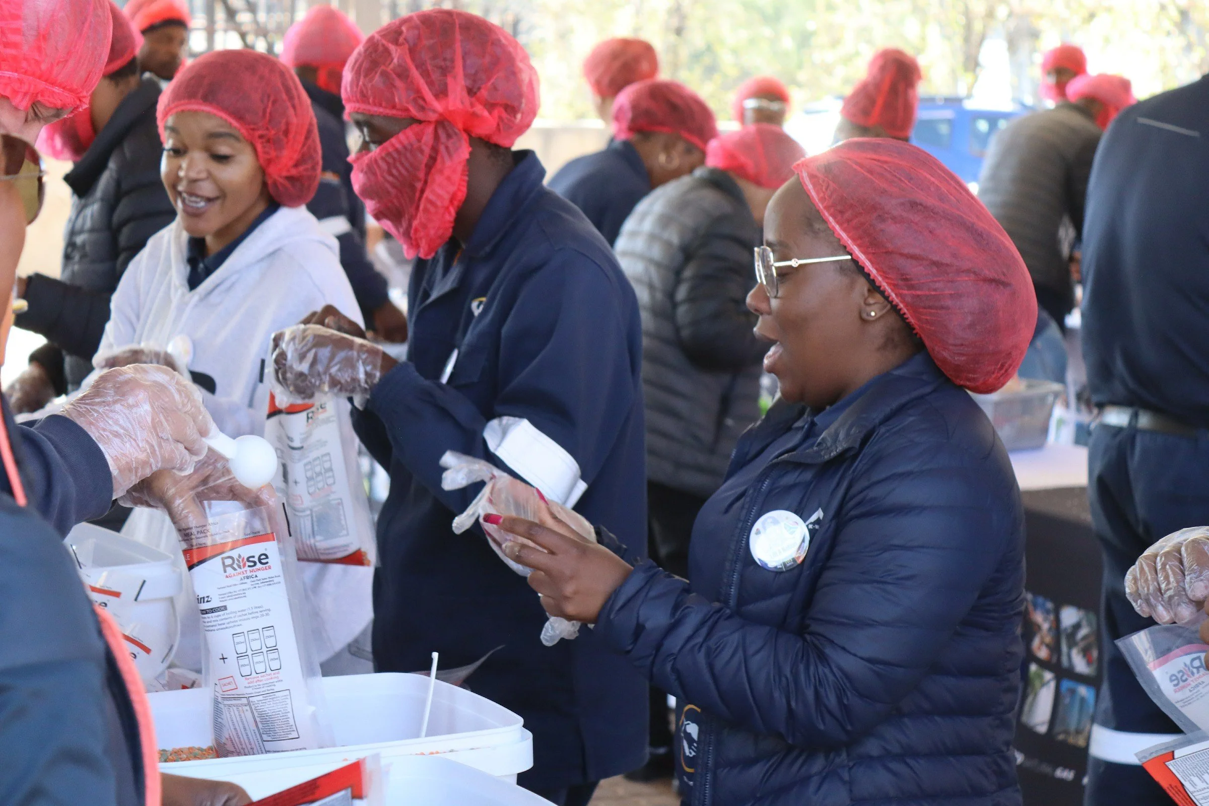 Group of people wearing red hairnet caps and gloves, working together at a table with food supplies, engaging in a charitable activity.