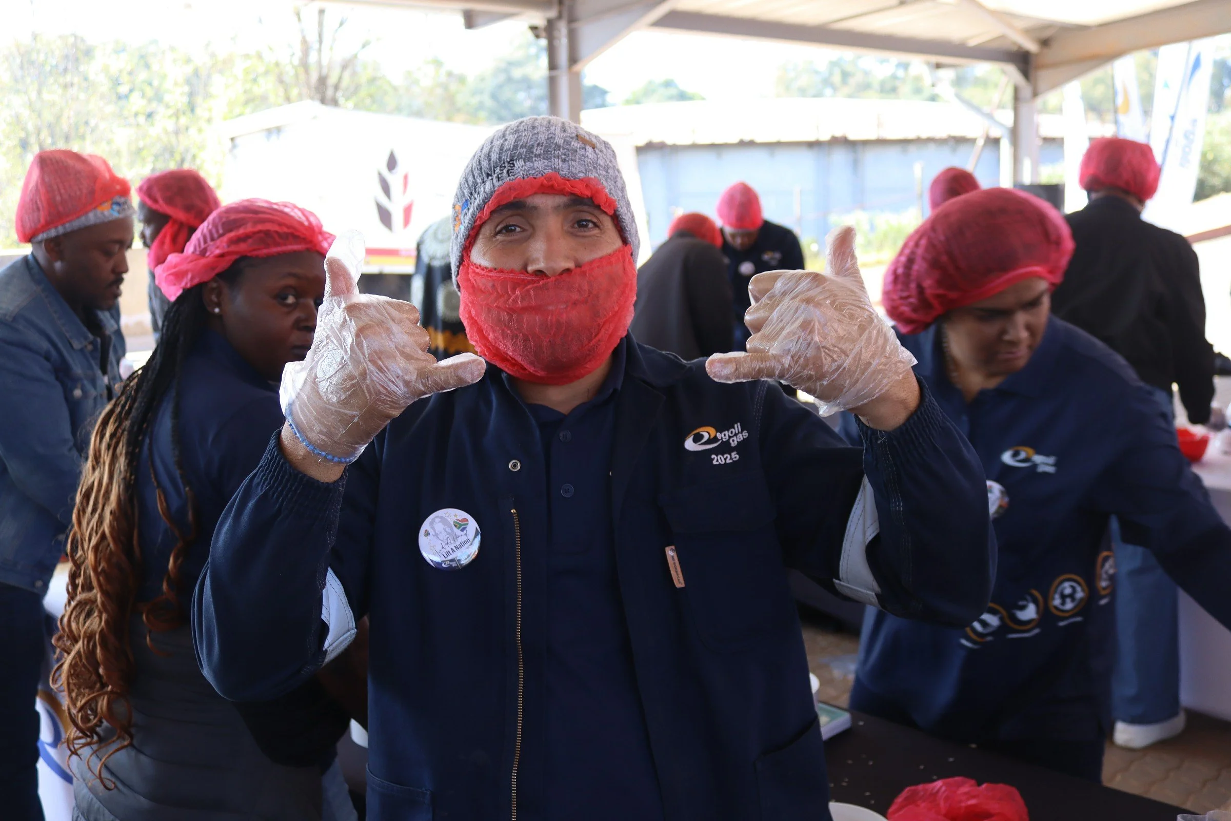 A group of people wearing hairnets and gloves at an outdoor event, with a woman in the foreground giving a thumbs-up gesture and smiling at the camera.