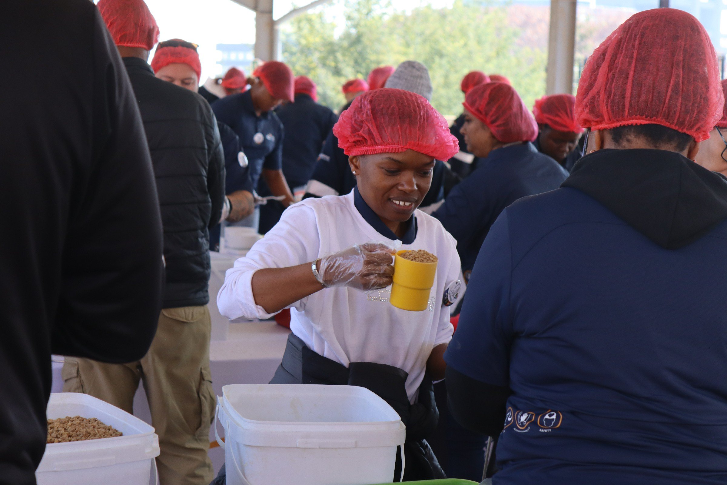 A diverse group of people wearing red hairnets and gloves, working together at a food packaging or distribution event with several large containers of food on the table.
