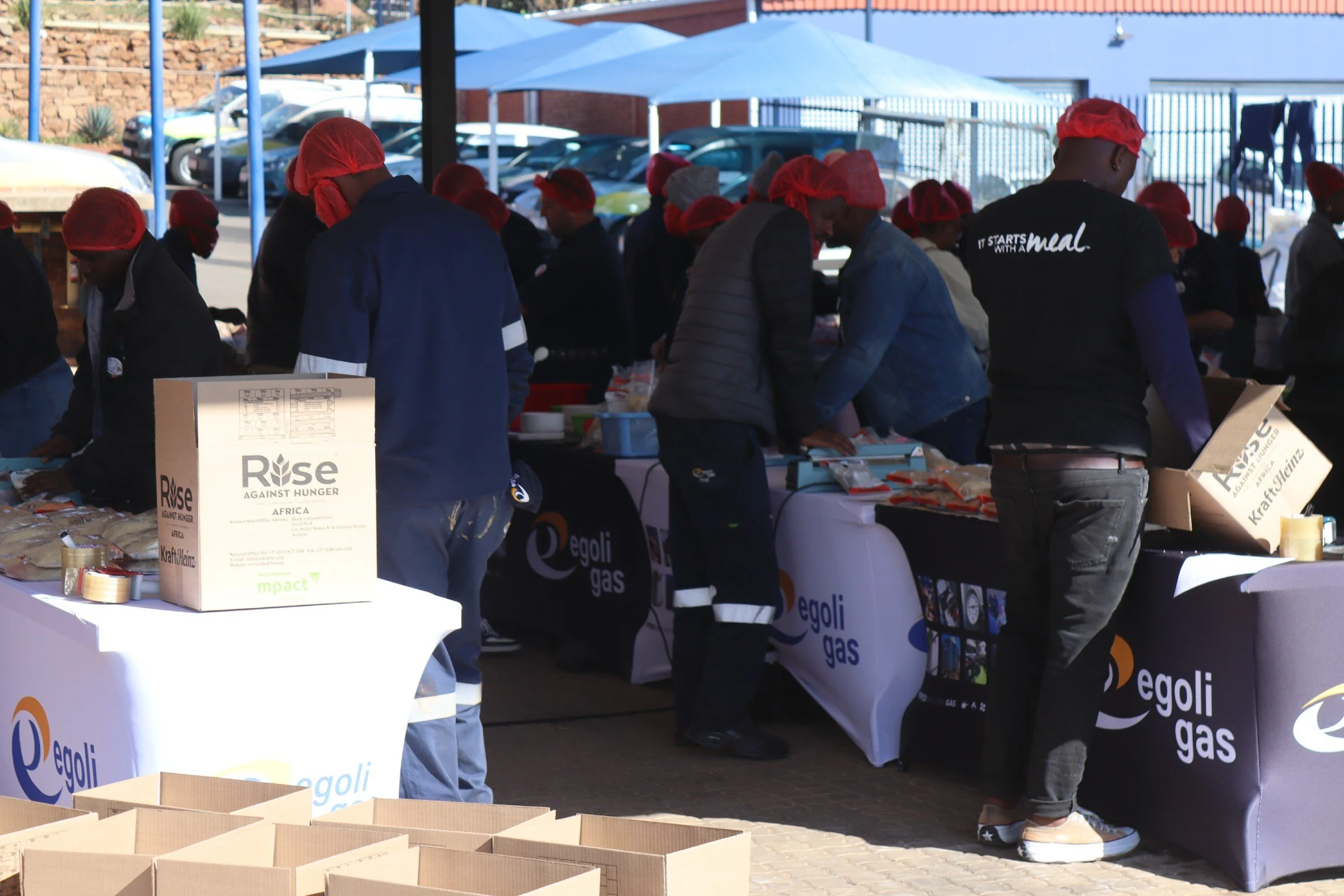 People wearing red hairnets working at a table with boxes and food items outdoors under large umbrellas, with cars parked in the background.