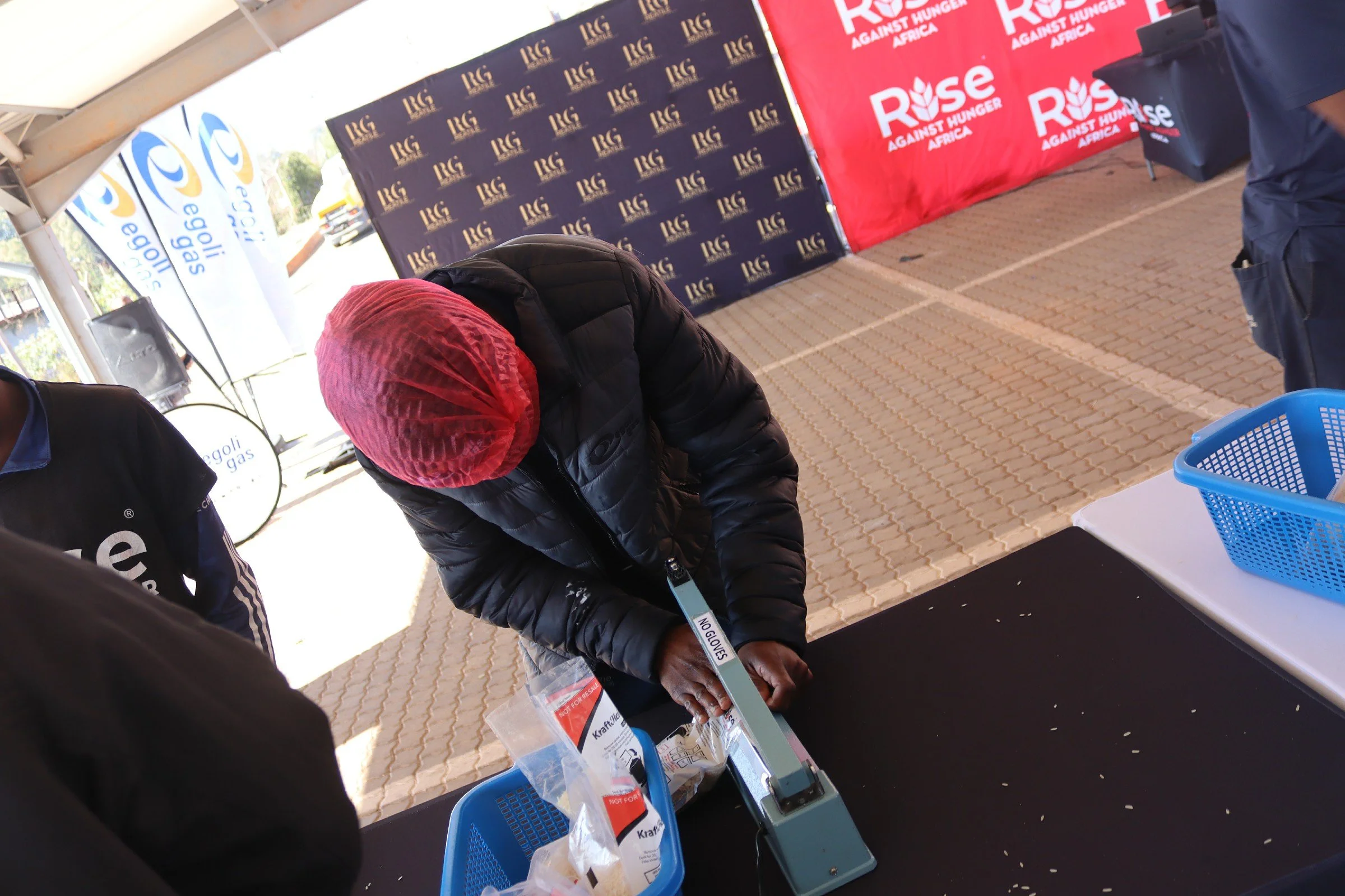 A person wearing a pink hair cover and a black puffy jacket is standing at a table, holding a clipboard with a sign that says 'NO GIVES'. There are banners in the background with logos and the words 'RISE AGAINST HUNGER AFRICA' and 'ELG'. A blue bask