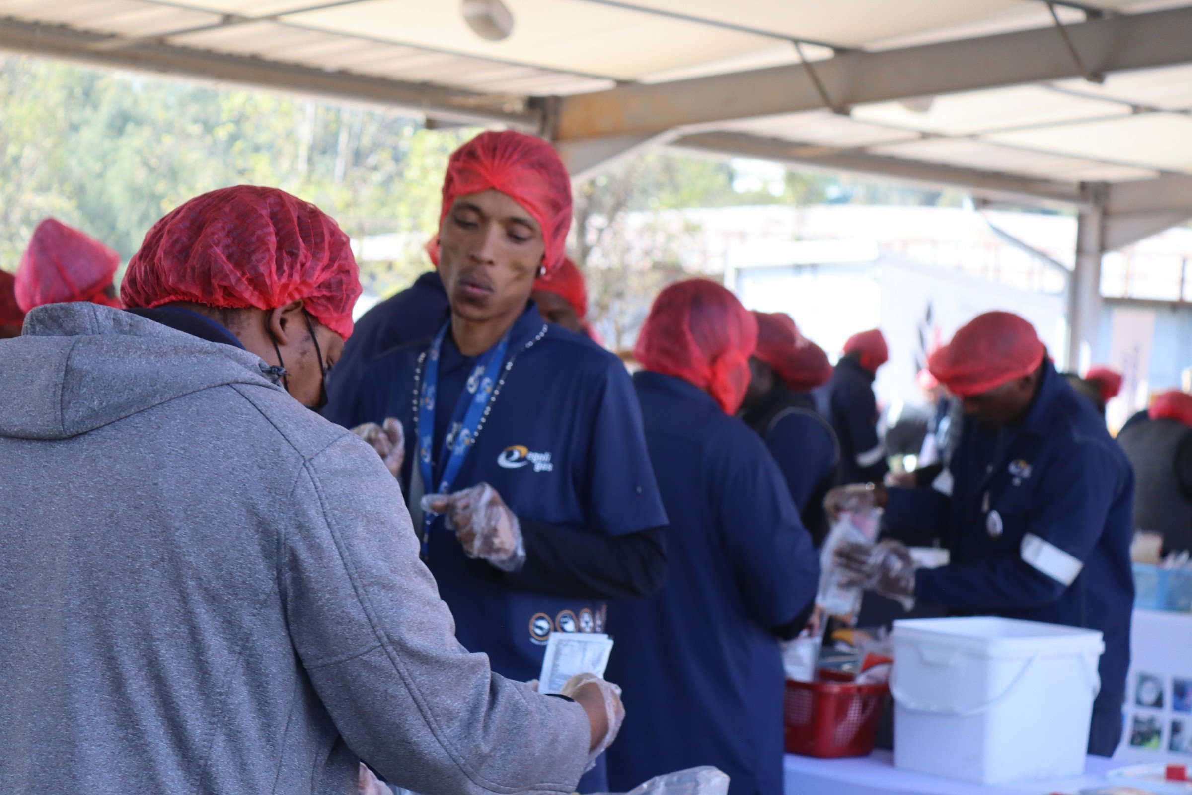 A group of people in a covered outdoor area, wearing red hairnets and dark jackets, are working together on a task, possibly packaging or preparing food, with trees and buildings visible in the background.