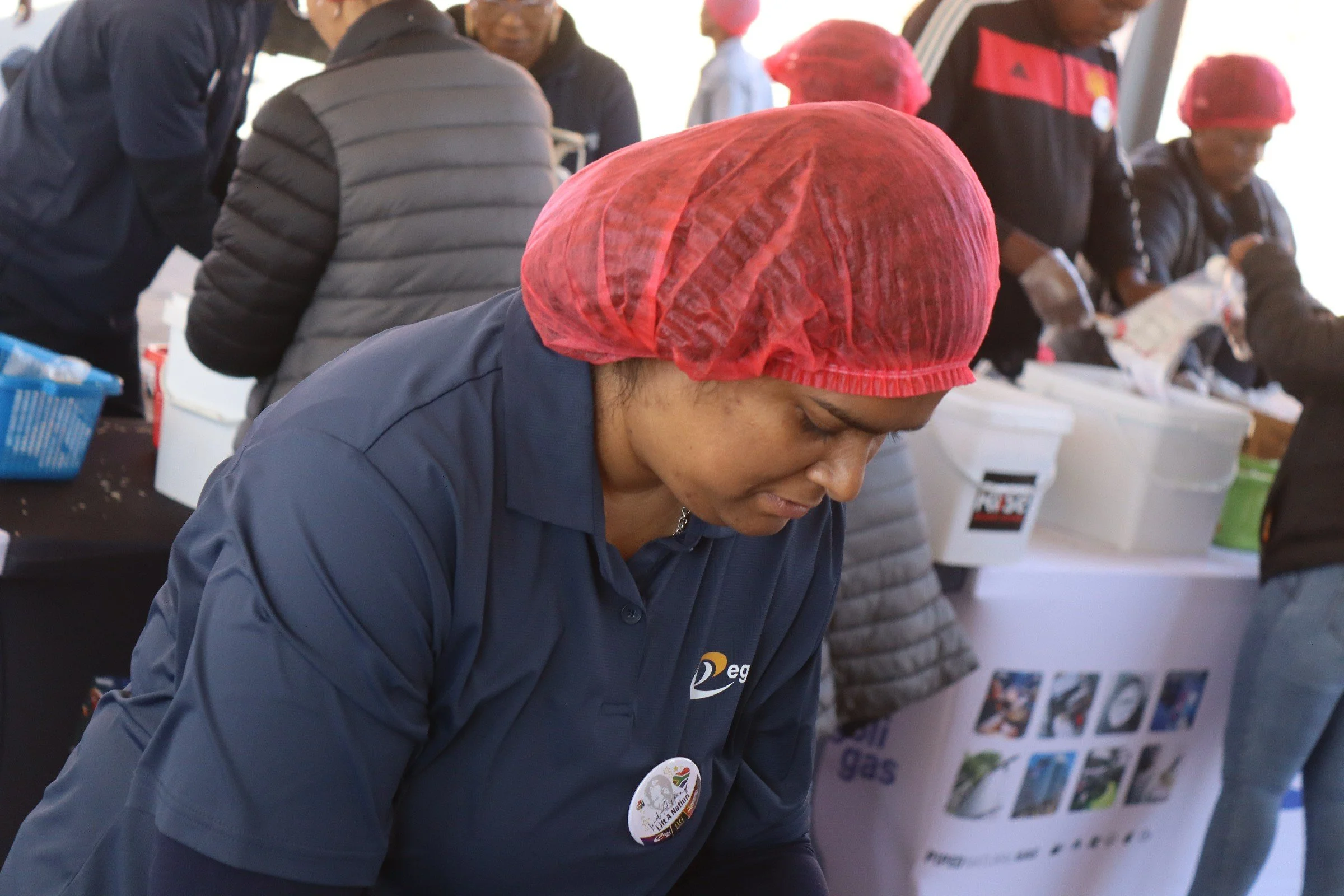 A woman wearing a red hairnet and a navy blue jacket with a badge, working at a table with several people, some of whom are wearing similar hairnets and jackets, in an indoor setting that appears to be a community event or food distribution.