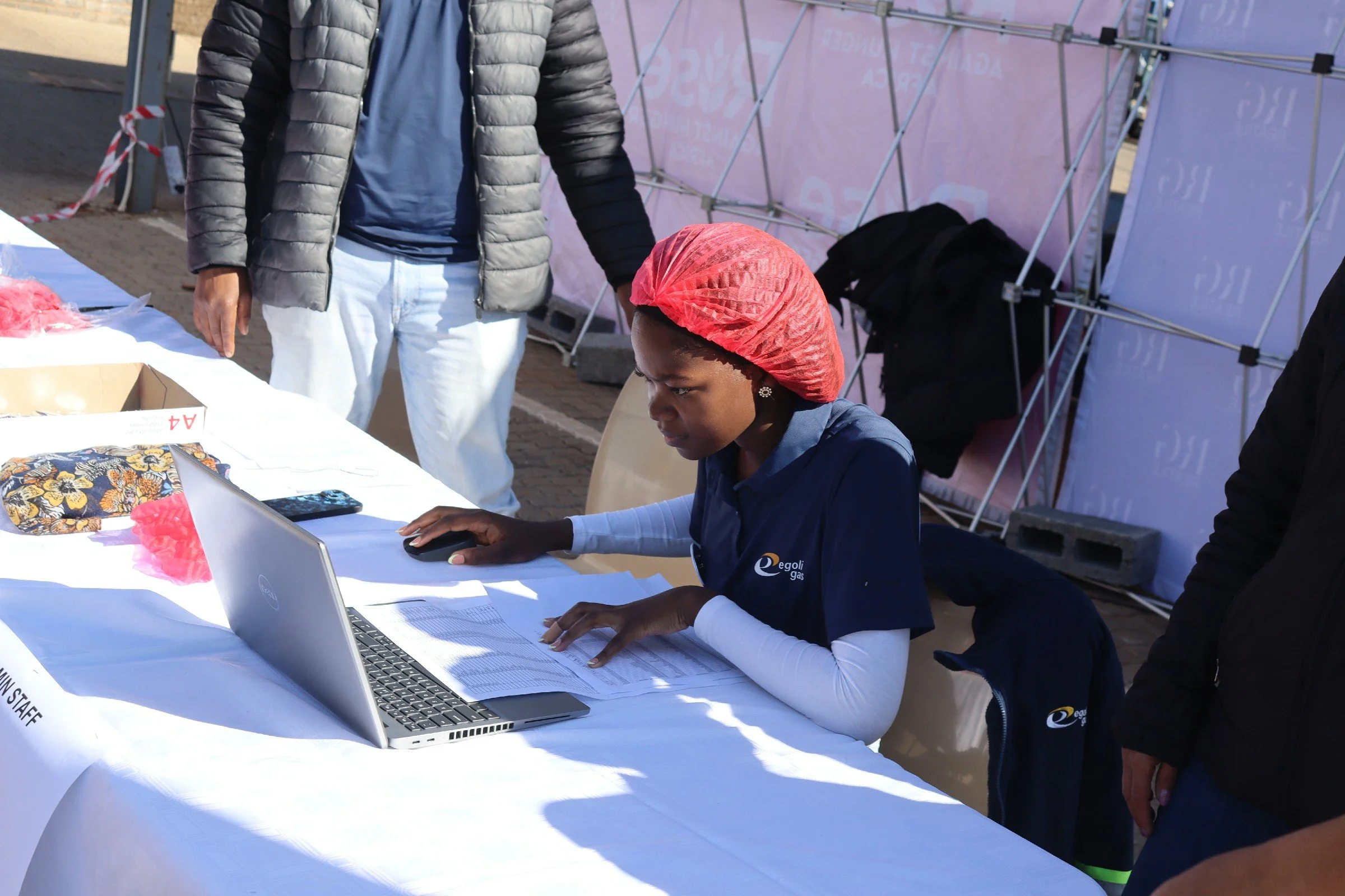 A woman sitting at a table with a laptop, papers, and a pen, using a computer mouse. She is wearing a navy blue shirt with a logo and a red head covering. A man in a puffy jacket stands nearby, and there are exhibit booths with pink walls and backpac