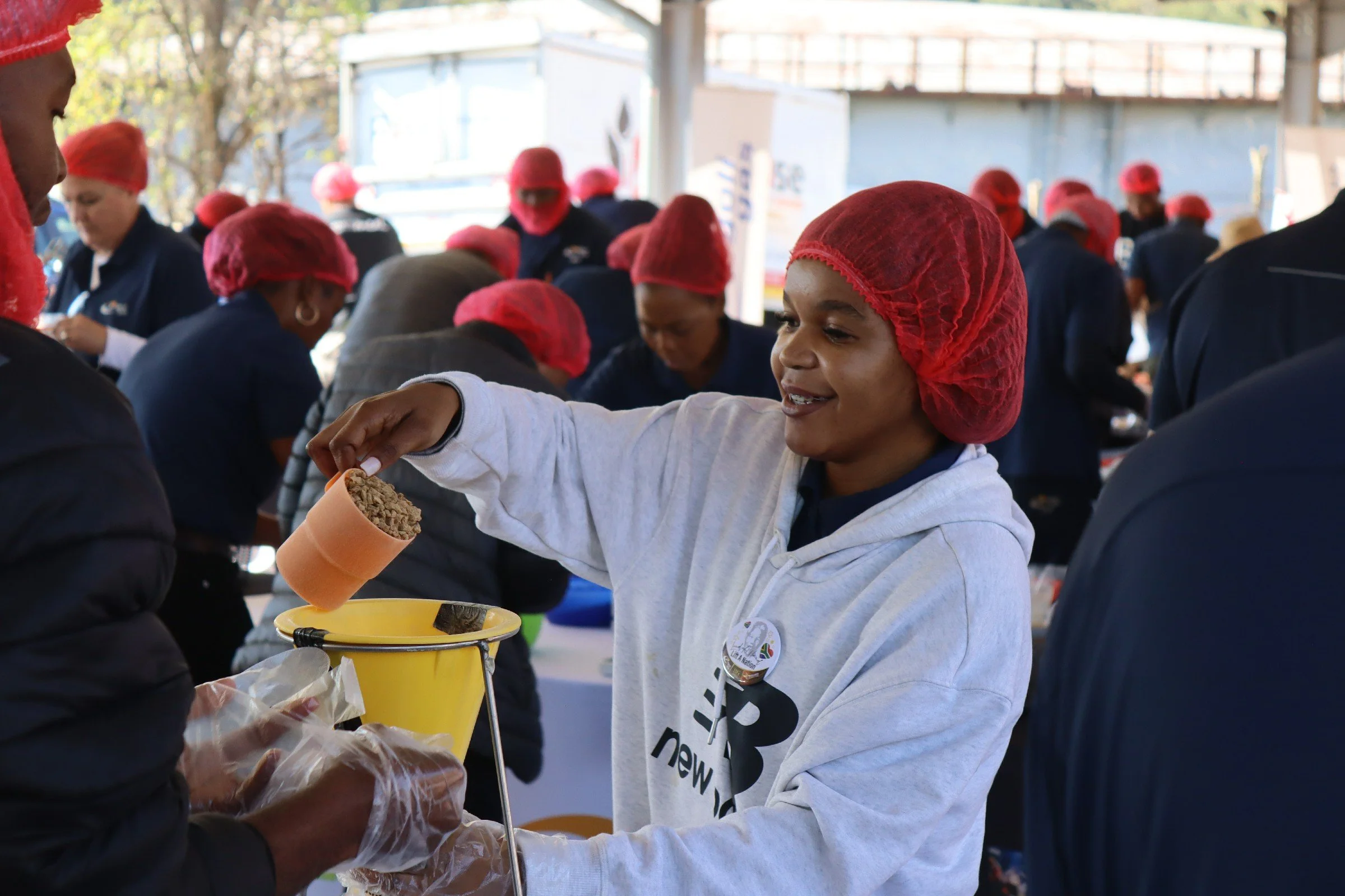 A young woman with a red hairnet receiving food from a person wearing black gloves at an outdoor service event with many people in red hairnets in the background.