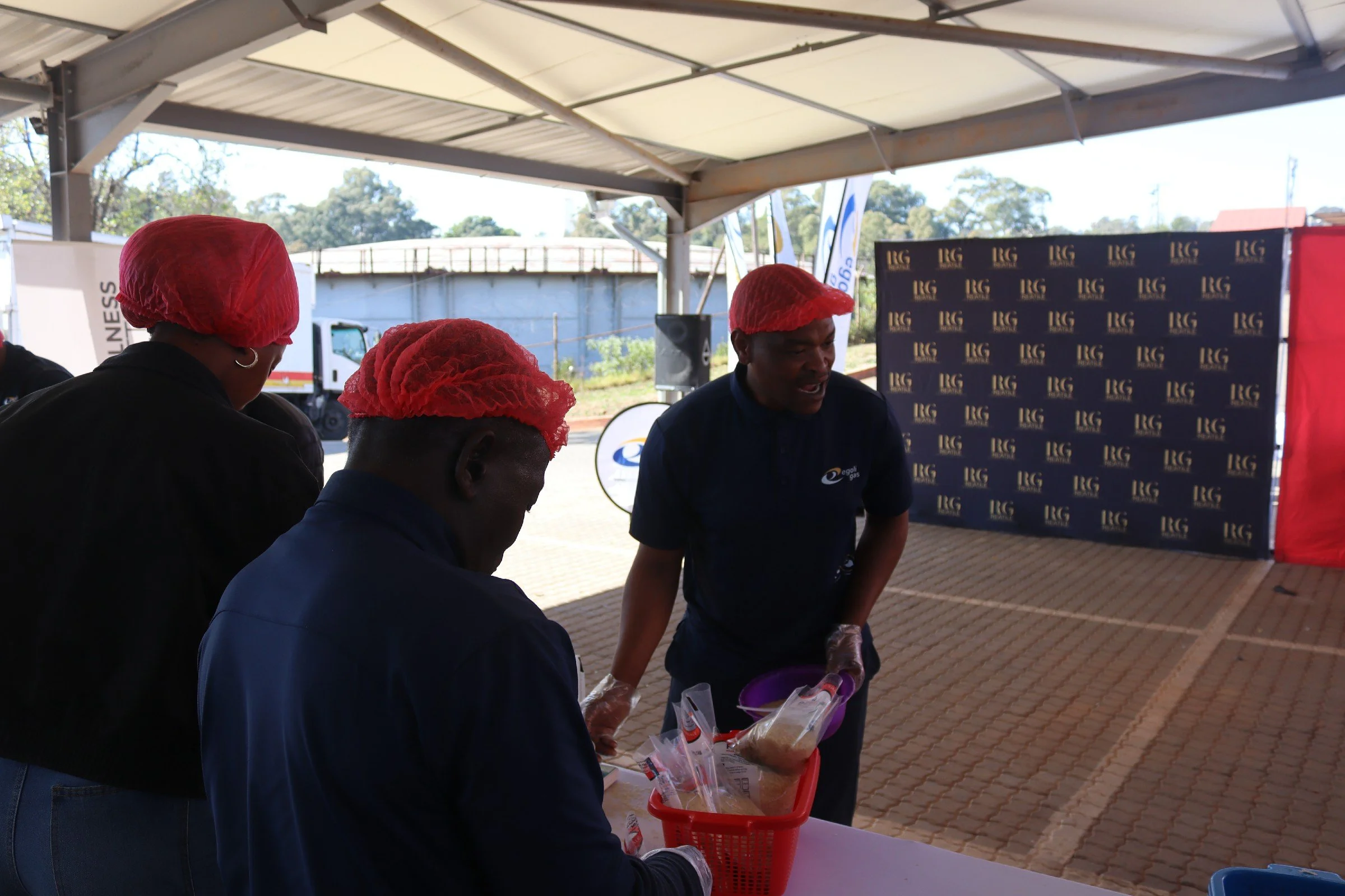 Three people wearing red hairnets serving food at an outdoor event under a tent.