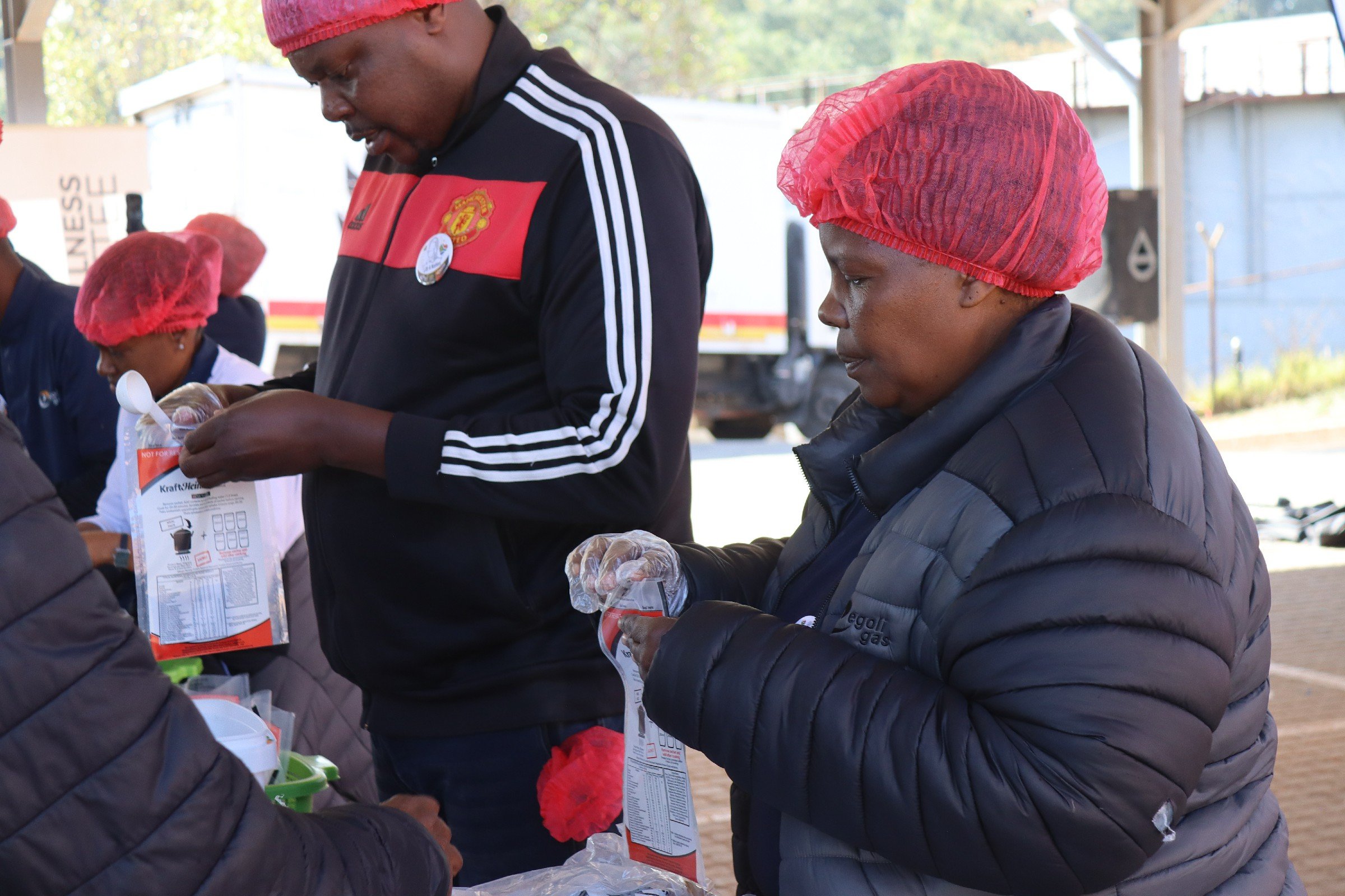 People wearing red hair covers and gloves, packing food bags at a community service event outdoors.
