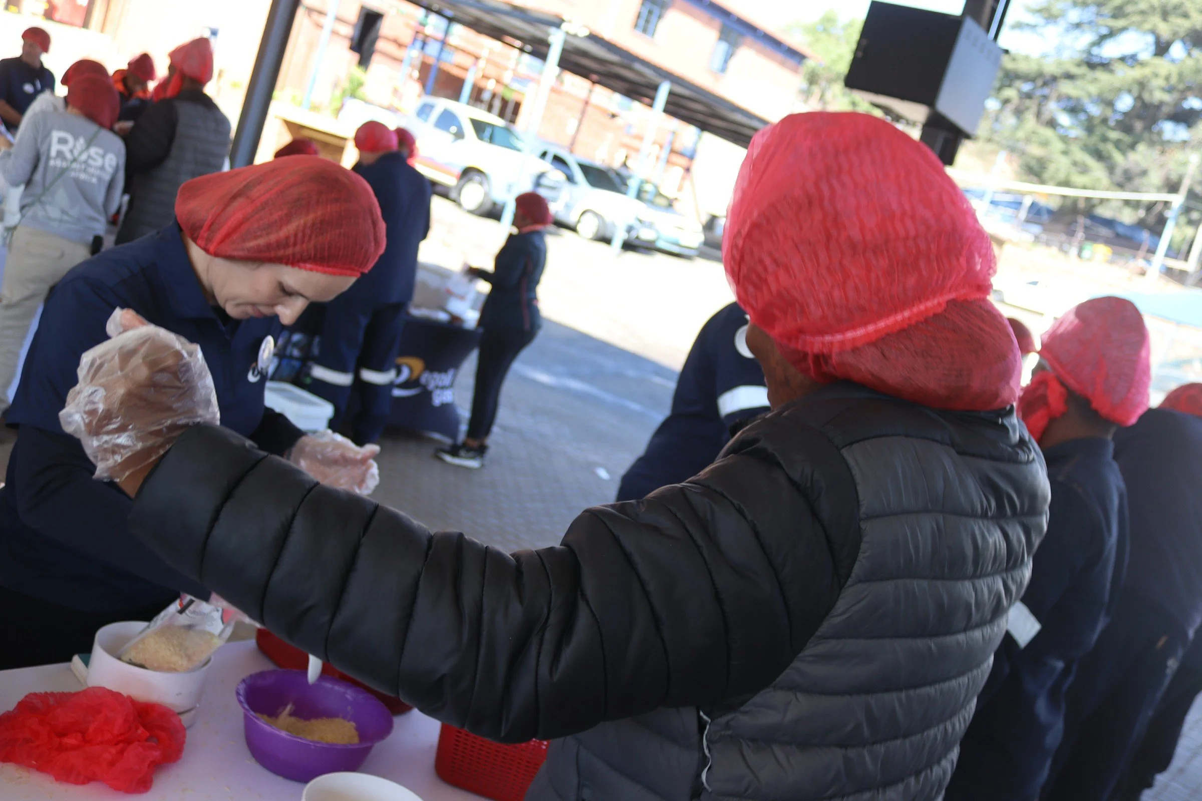 People wearing red head coverings gather outdoors around a table with bowls of food, likely celebrating a festive event.