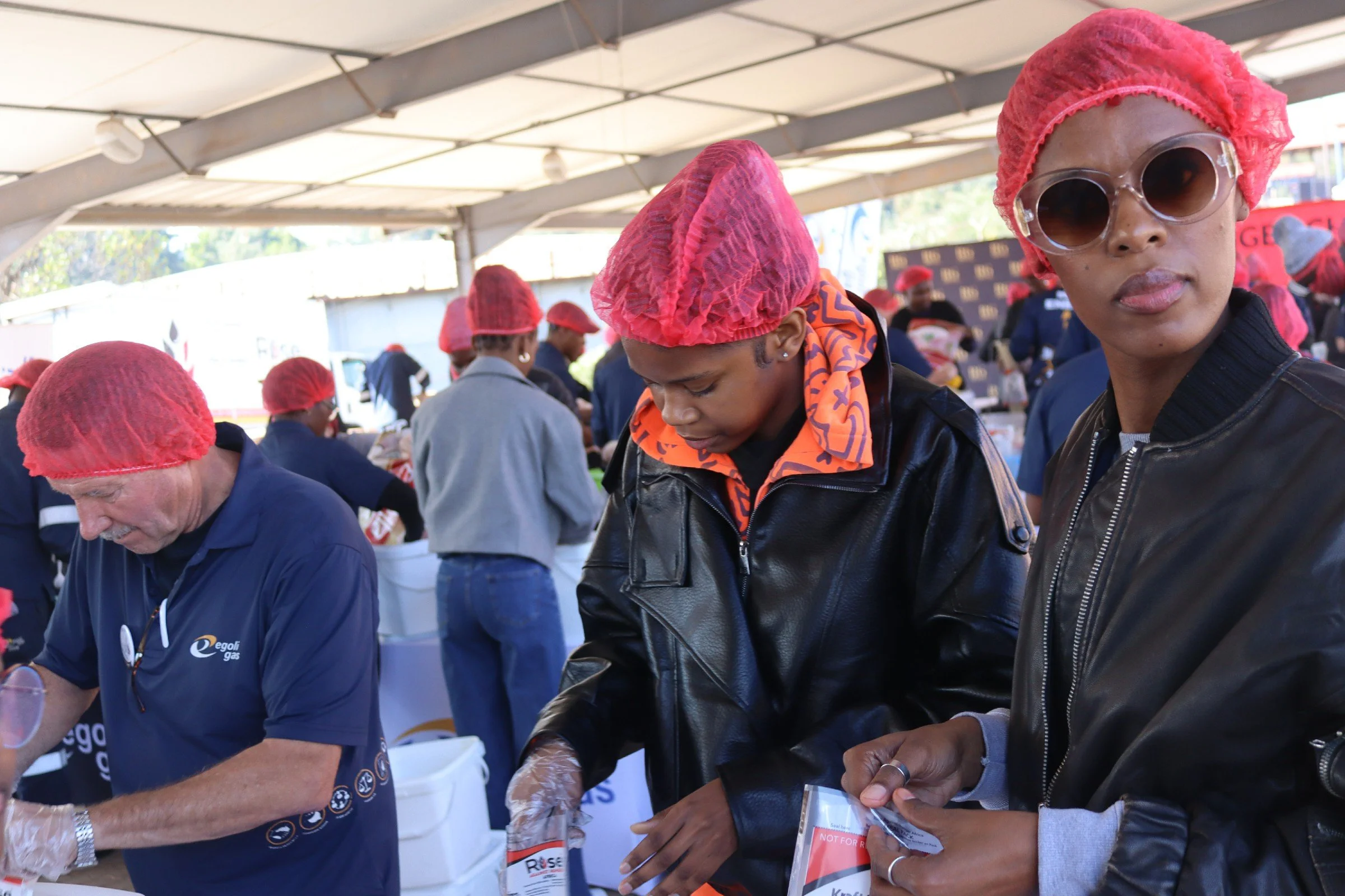 Group of people wearing red hairnets, some with sunglasses, participating in an outdoor event under a large canopy tent, serving or preparing food.