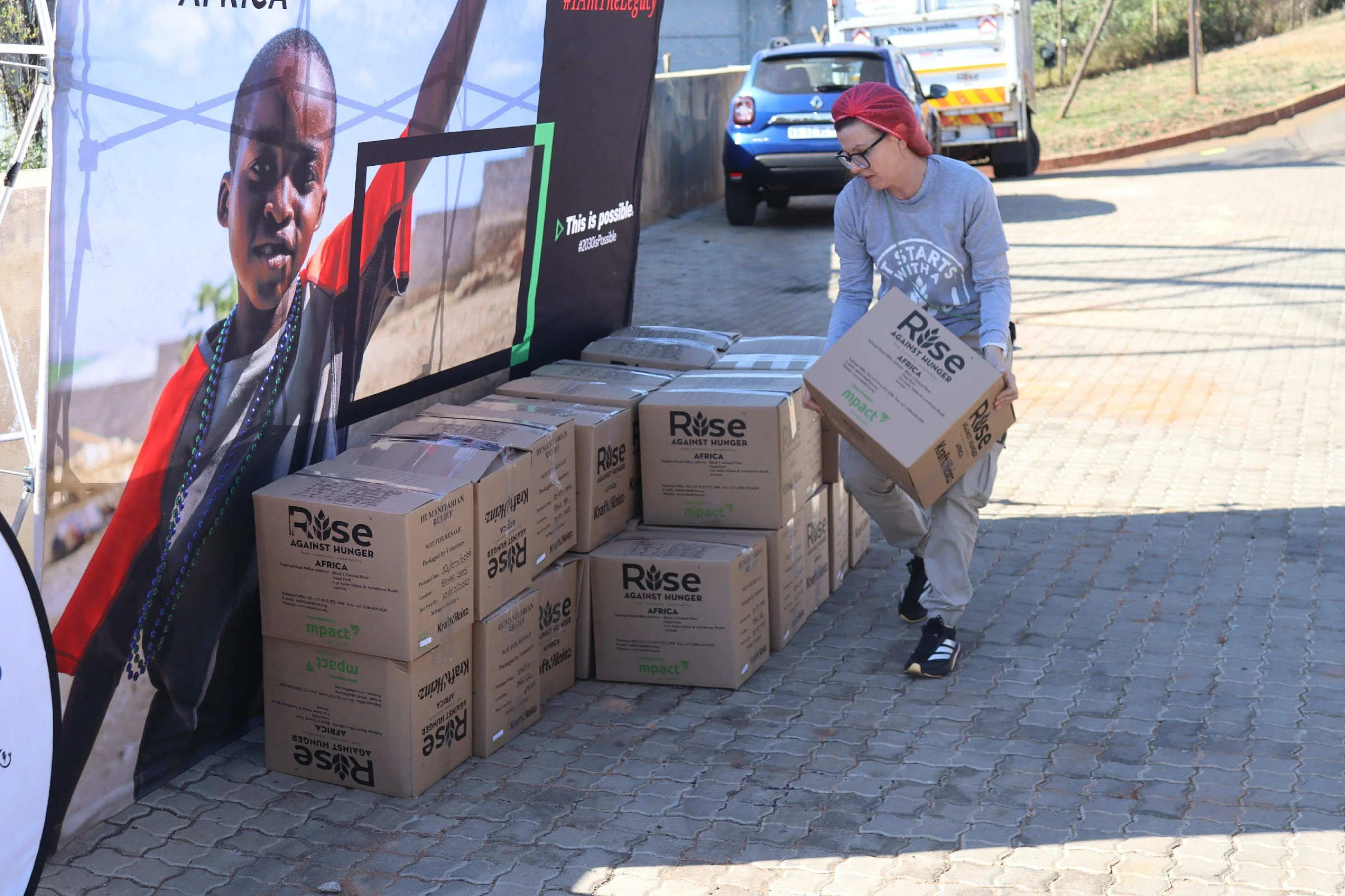 A woman with red hair, black glasses, wearing a grey long-sleeve shirt, black pants, and sneakers, is stacking boxes labeled 'Rise Against Hunger' at an outdoor event. There are many boxes on the ground, and a large banner with a child's face and the