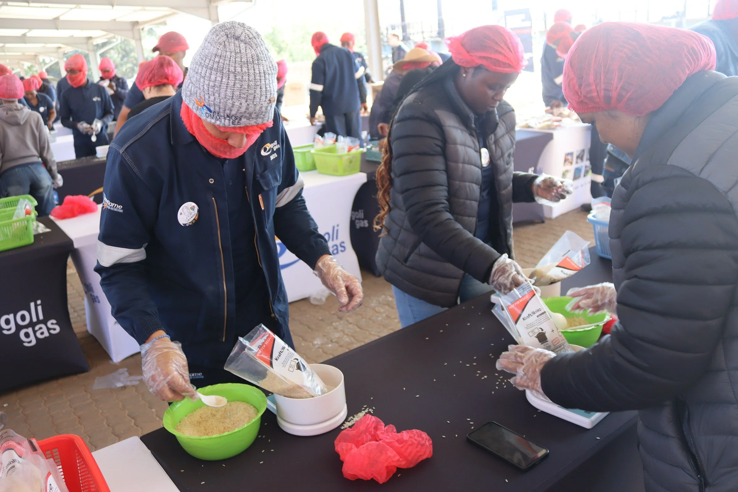 People wearing gloves and hair coverings working at a table with food ingredients during a cooking event or workshop.