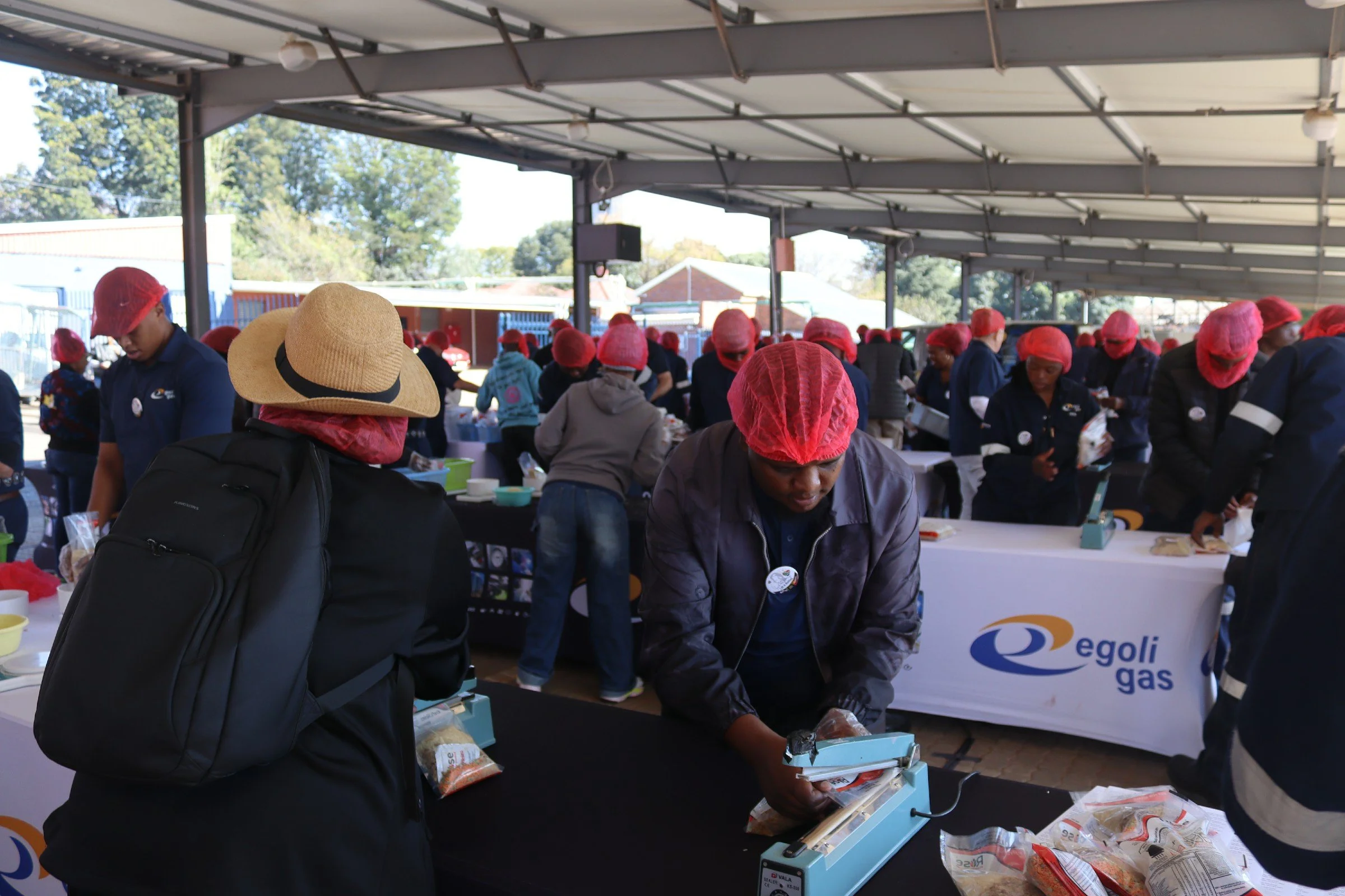 A group of people wearing red hairnets and dark clothing at an outdoor event, some are working at tables with food products, under a large canopy with trees and a building in the background.