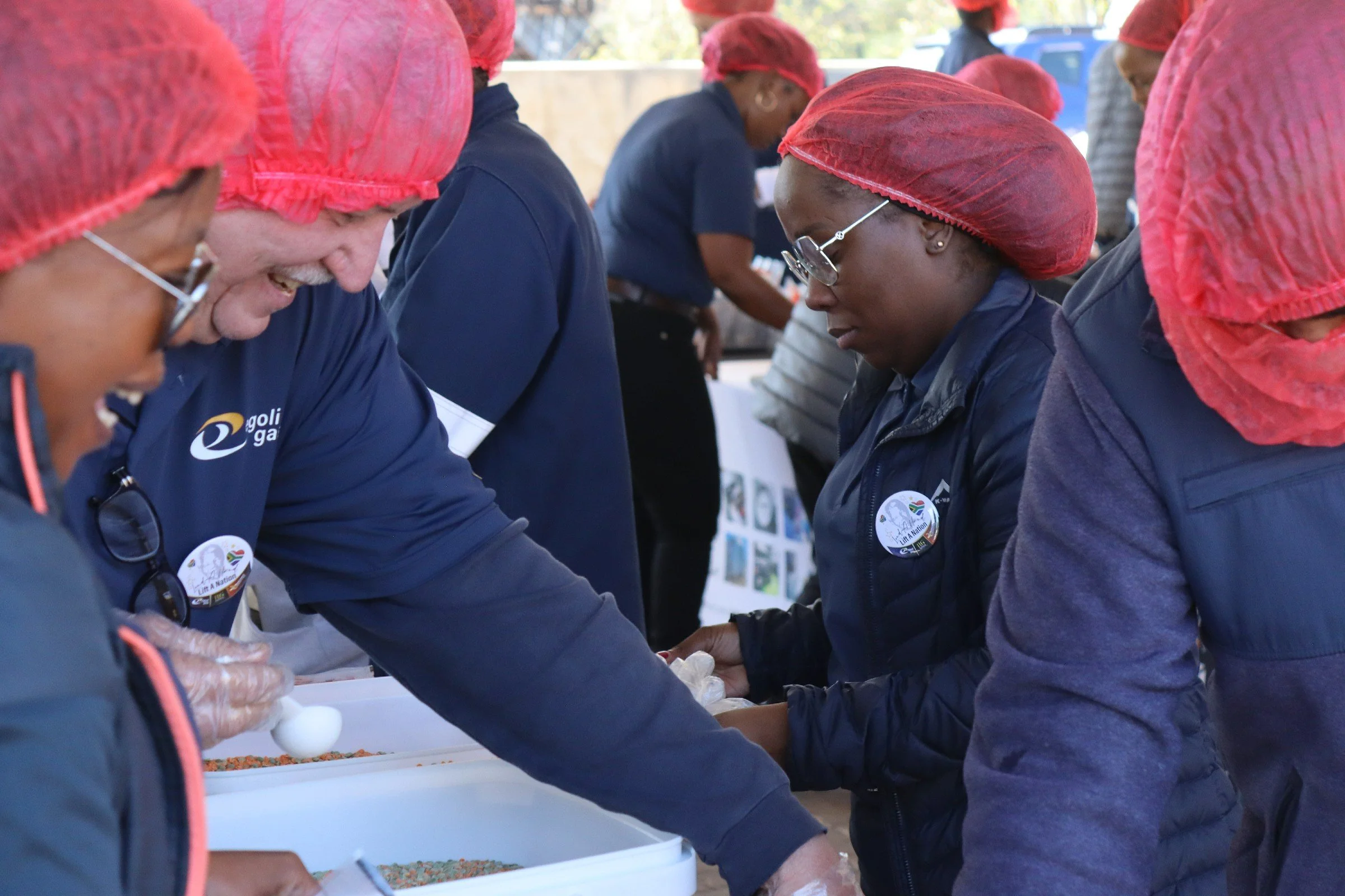 People wearing red hair covers and dark clothing participating in an outdoor event, working with small objects on a table.