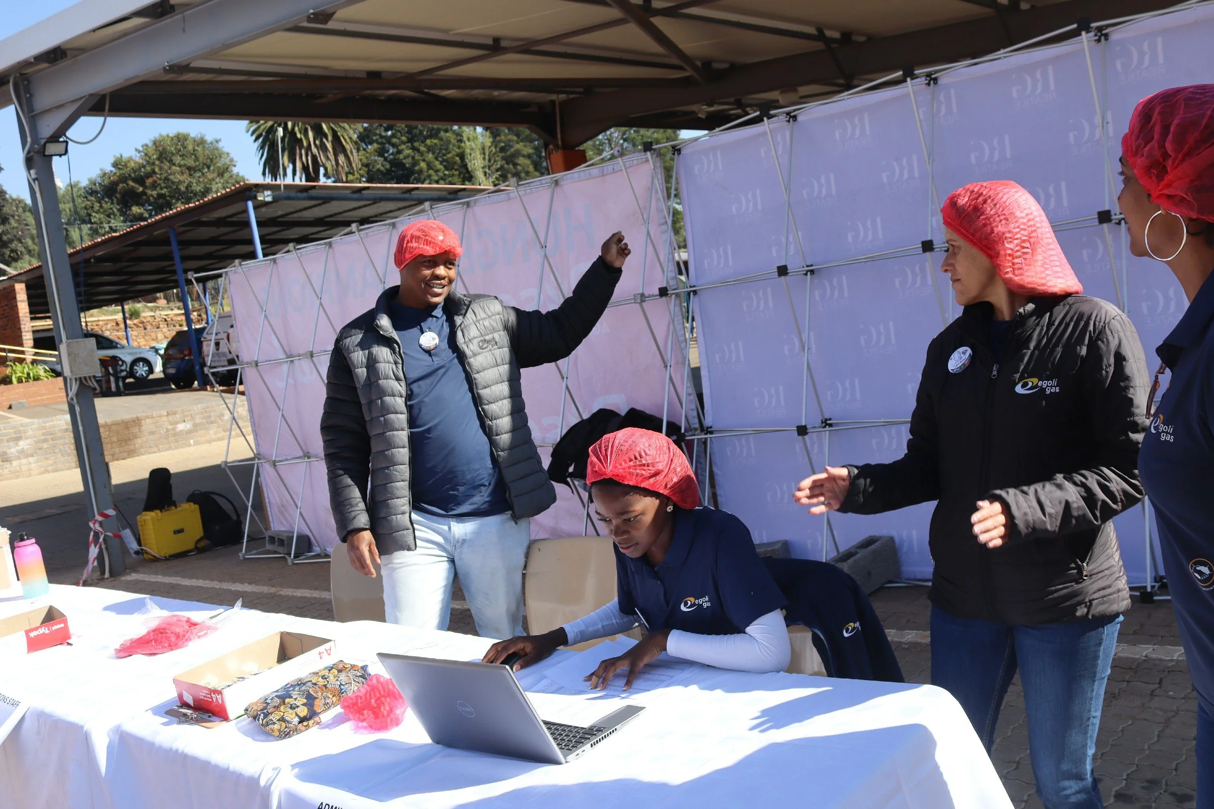 People at an outdoor event or booth, some wearing red hair caps, one person speaking or explaining, others working at a table with a laptop and various items, under a canopy with a backdrop that has a logo or text.