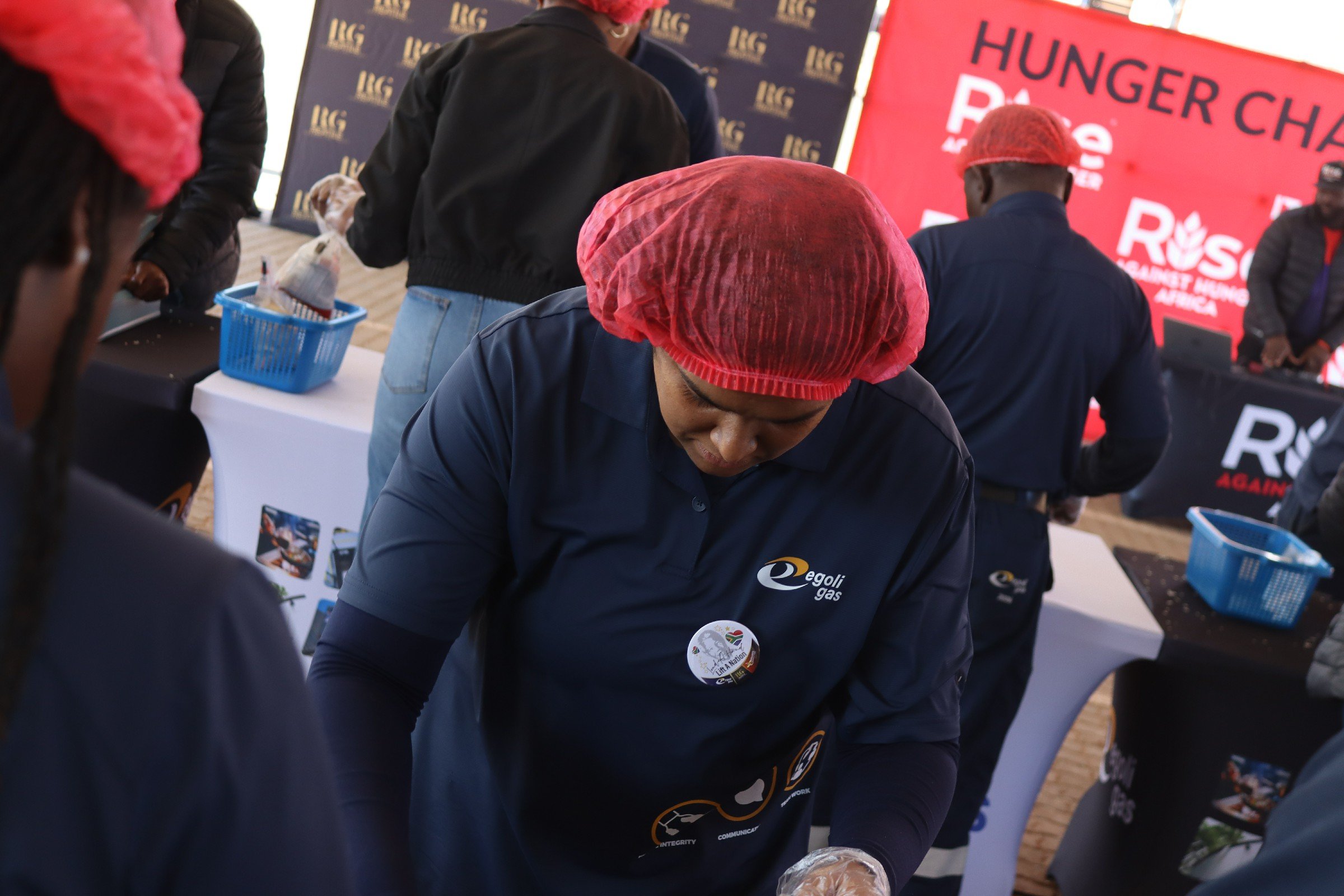 People wearing red hairnets and dark shirts serving food at a Hunger Pledge event with a red and black banner in the background.