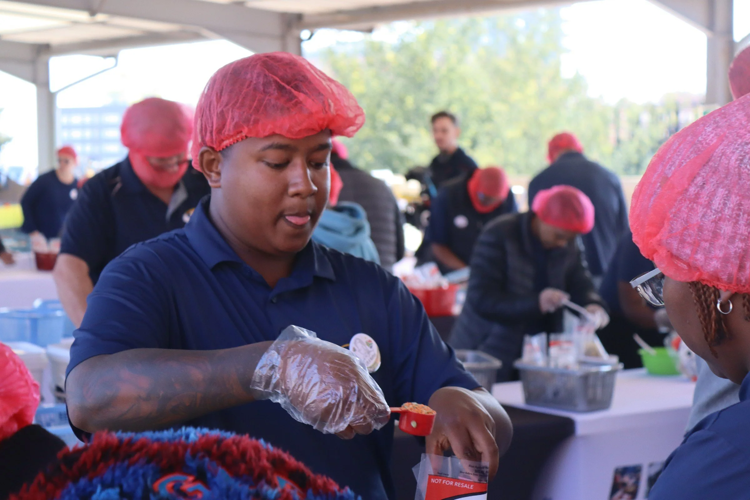 Young people serving food at a community event under a canopy, wearing red hairnets and dark clothing.