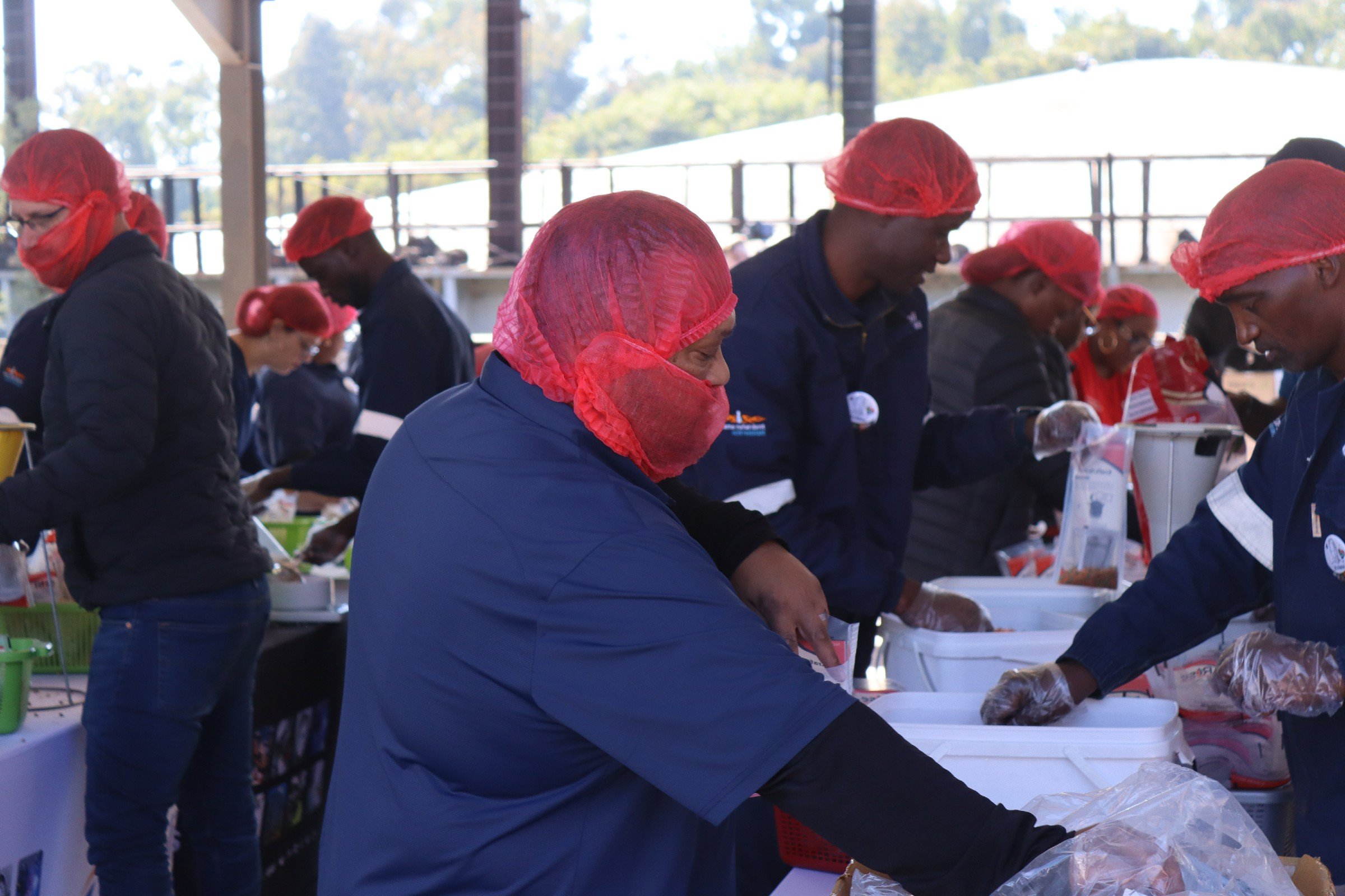 People working at a food processing or packaging event, wearing red hairnets and gloves, standing near white tables under a large open structure with an outdoor background.