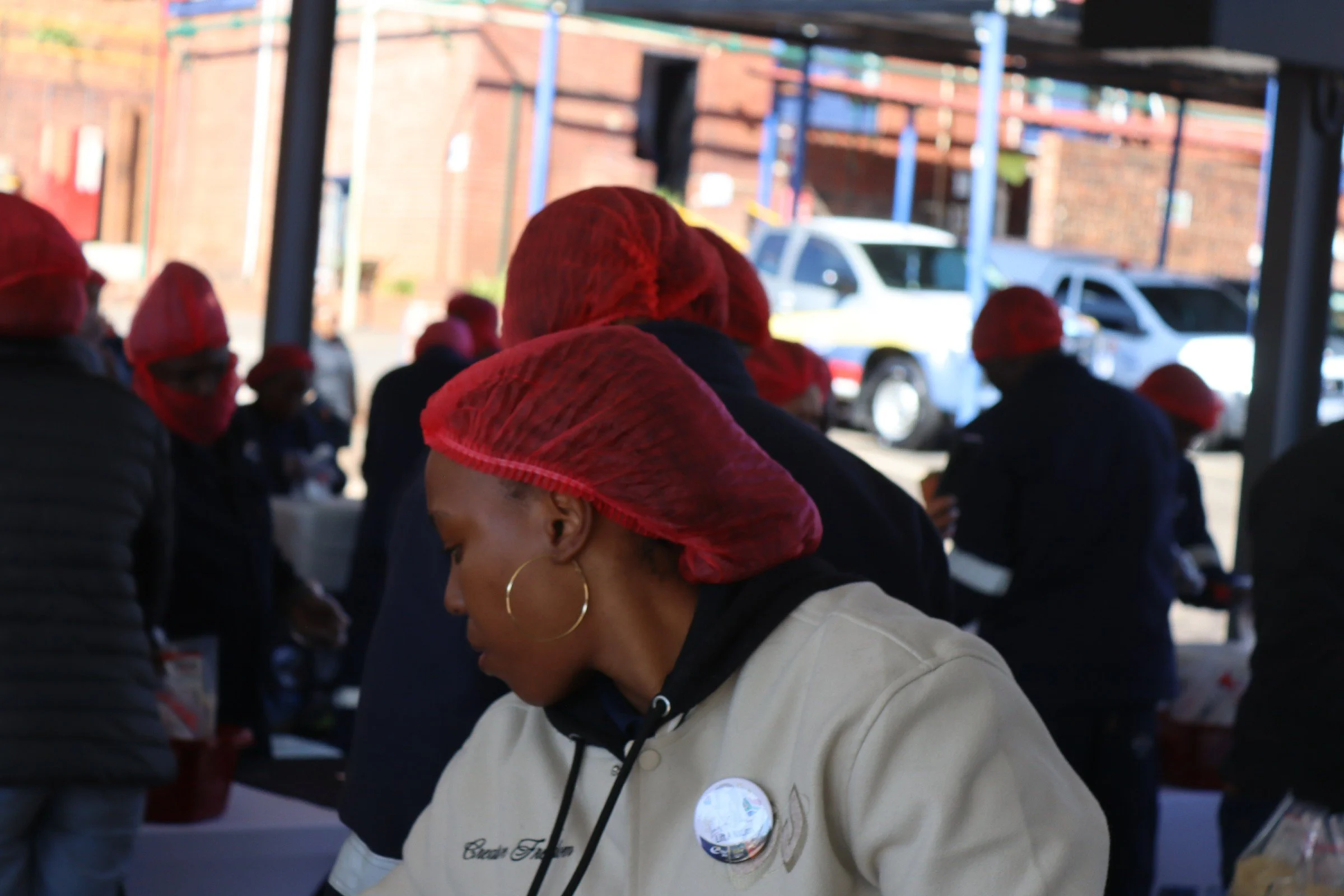 A group of people wearing red hair coverings gathered outside, with a brick building and parked vehicles in the background.