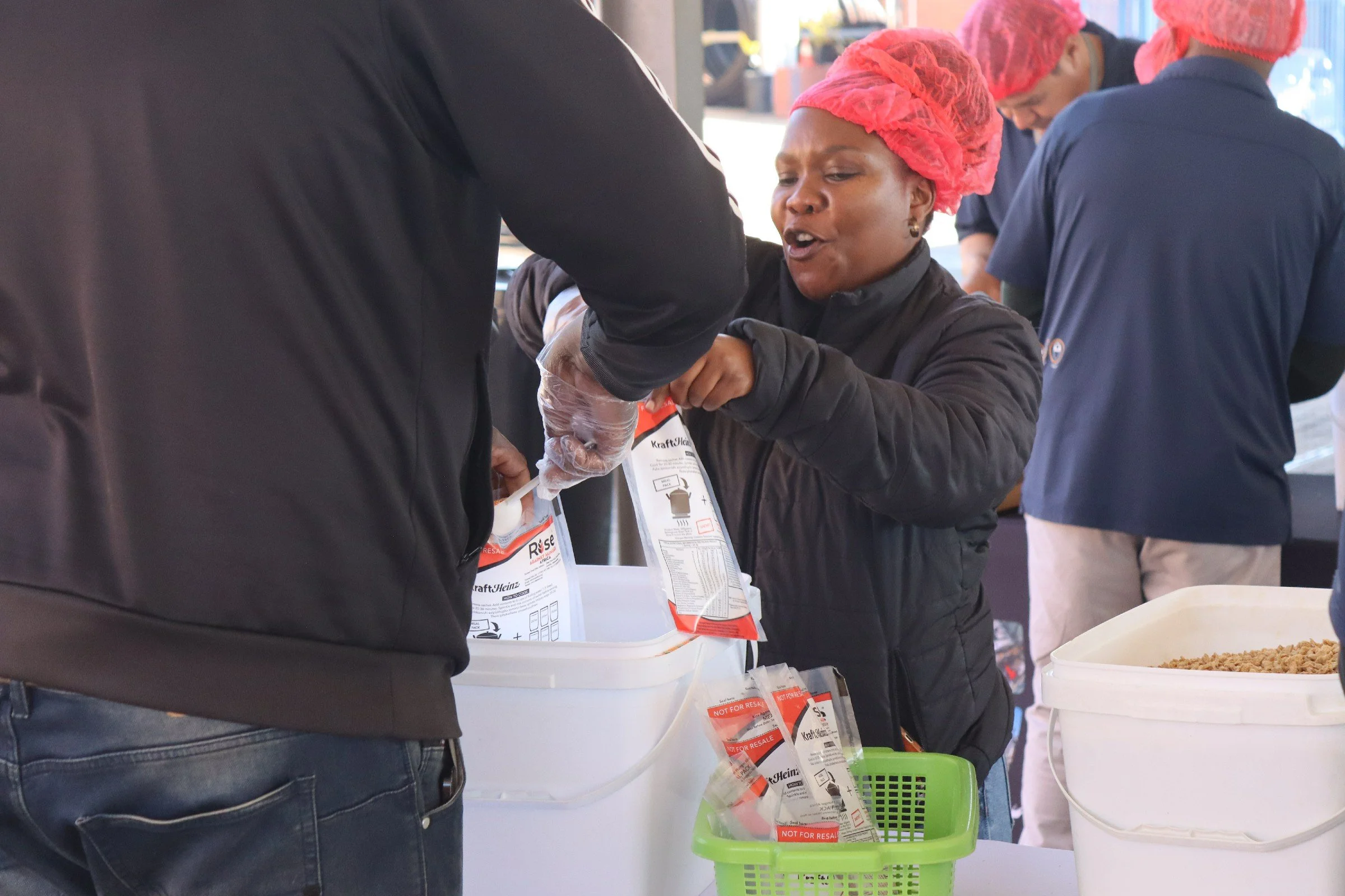 A woman with a pink hairnet and black jacket is volunteering at a community food distribution. She is handing a bag of food to a person, and there are other volunteers in pink hairnets and blue uniforms in the background.