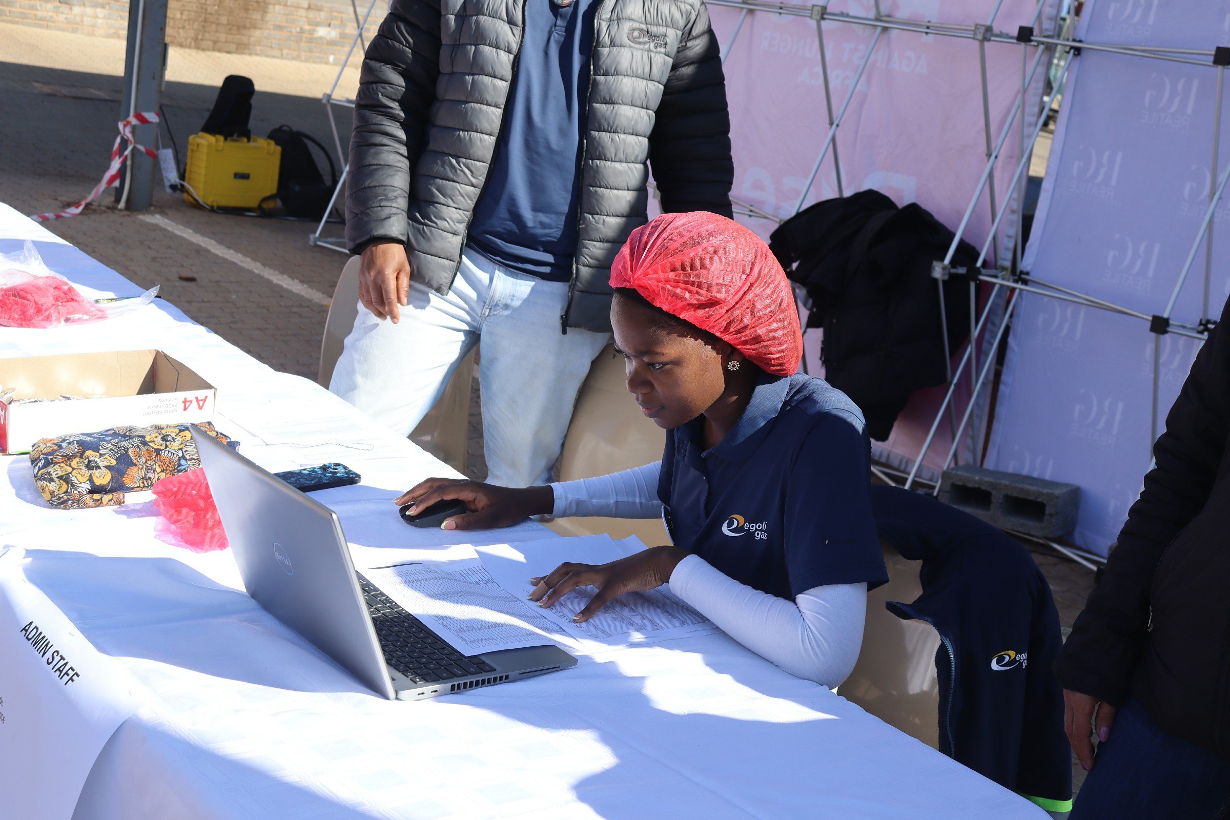 A person sitting at a table working on a laptop, with another person standing nearby. The table has various items including a cloth, a box, and paperwork. The background suggests an outdoor event with tents and equipment.