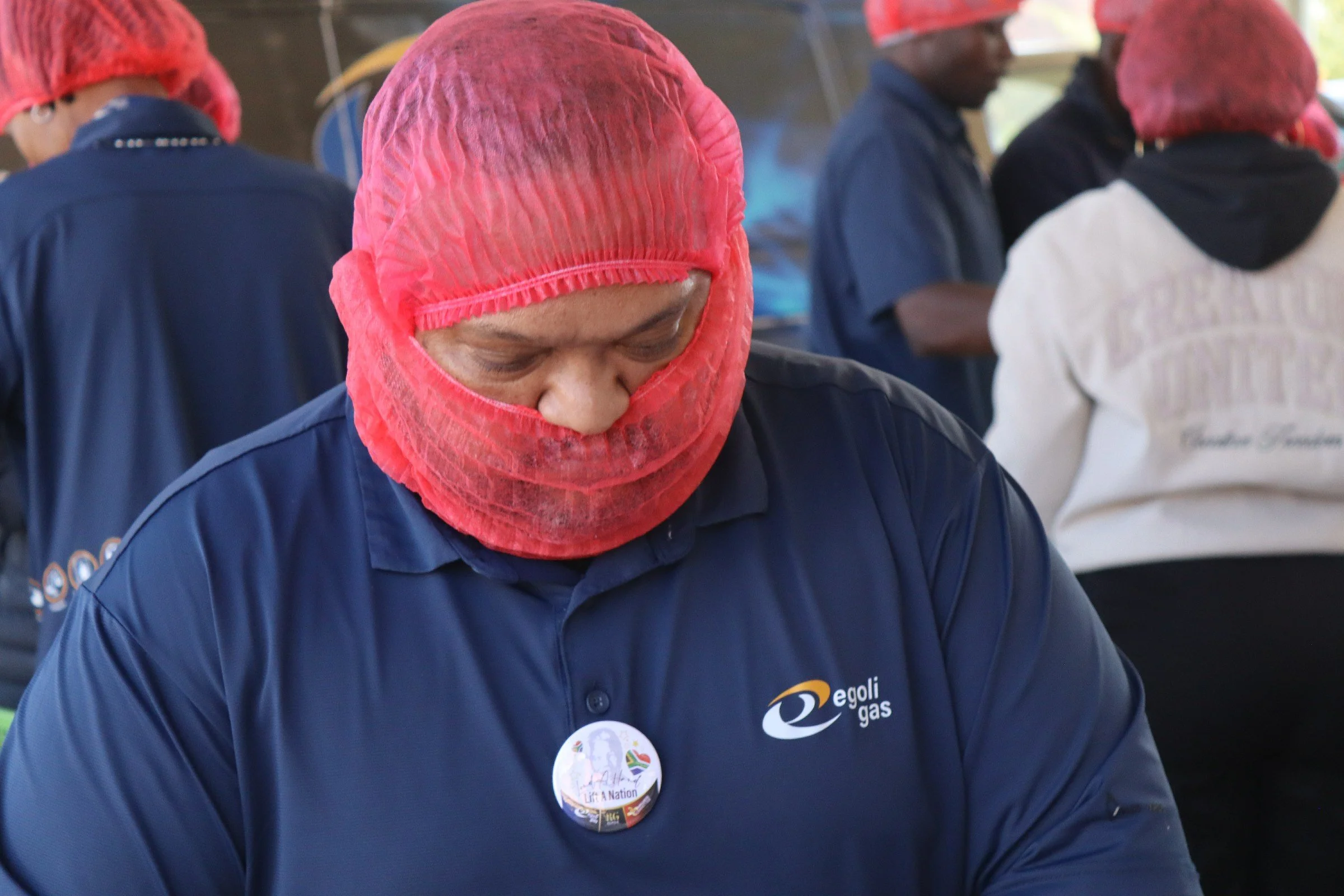 A person wearing a red hairnet and a dark blue shirt with a logo, looking down. Other people in similar attire, also wearing red hairnets, are in the background.