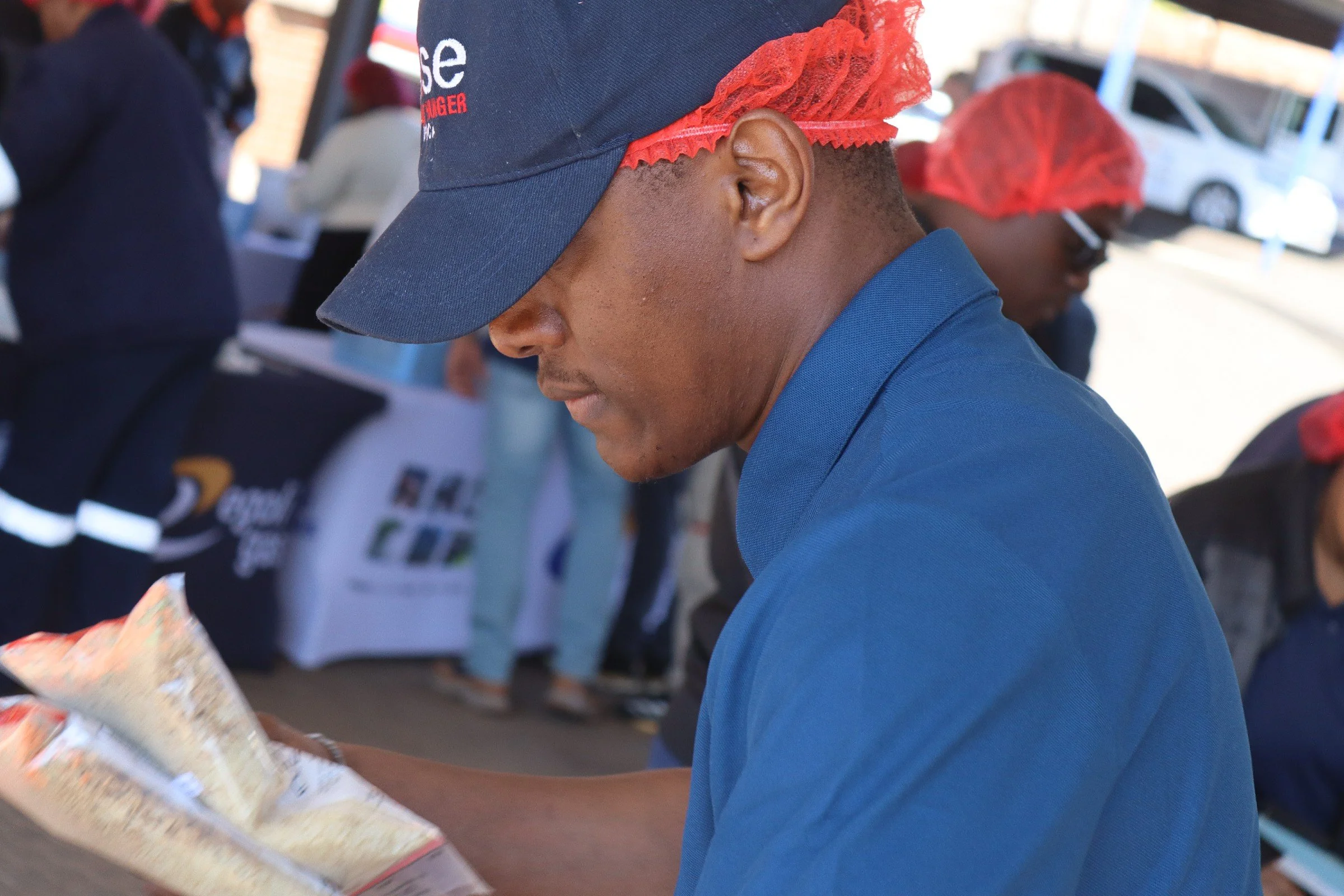 A person wearing a navy baseball cap with red trim and a red hairnet, dressed in a blue shirt, is handing money to someone at a vendor stand. Other people are visible in the background, some with similar hairnets, and there are parked cars and a bric