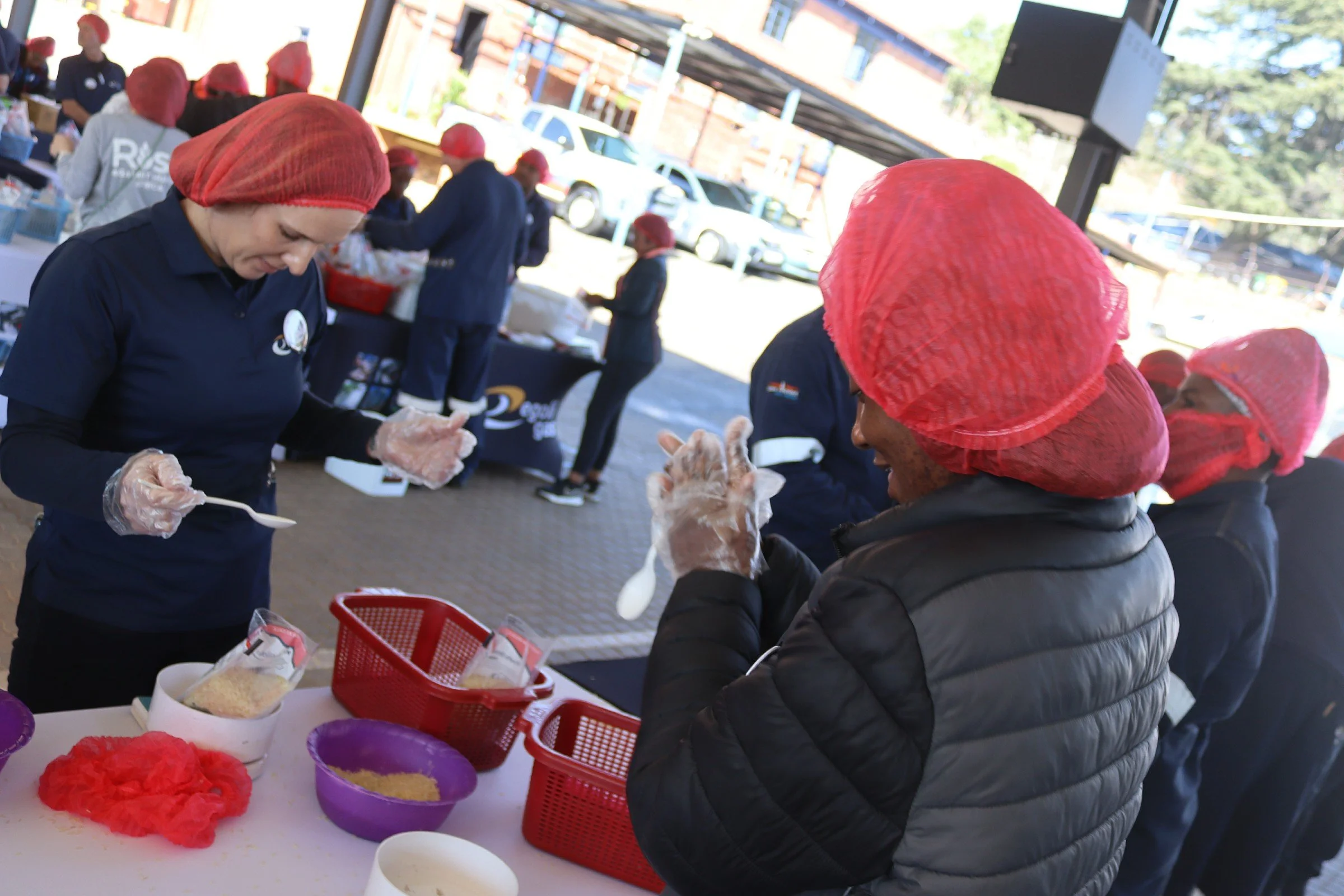 People wearing red hairnets and black jackets working at a table with colorful craft supplies, possibly making crafts or decorations, outdoors under a shelter.