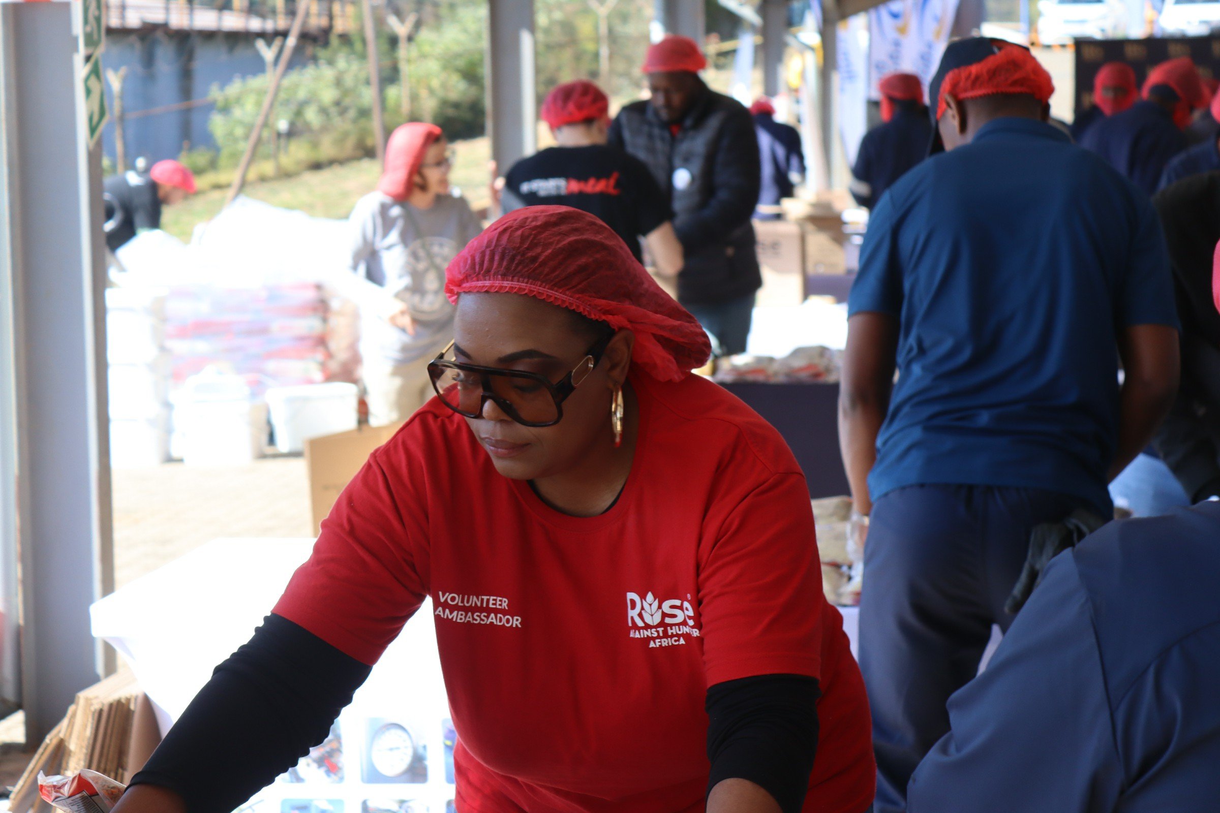 A woman wearing a red shirt, glasses, and a red hairnet assisting at a volunteer event, surrounded by other volunteers in red hairnets and blue shirts working together outdoors under a shelter.