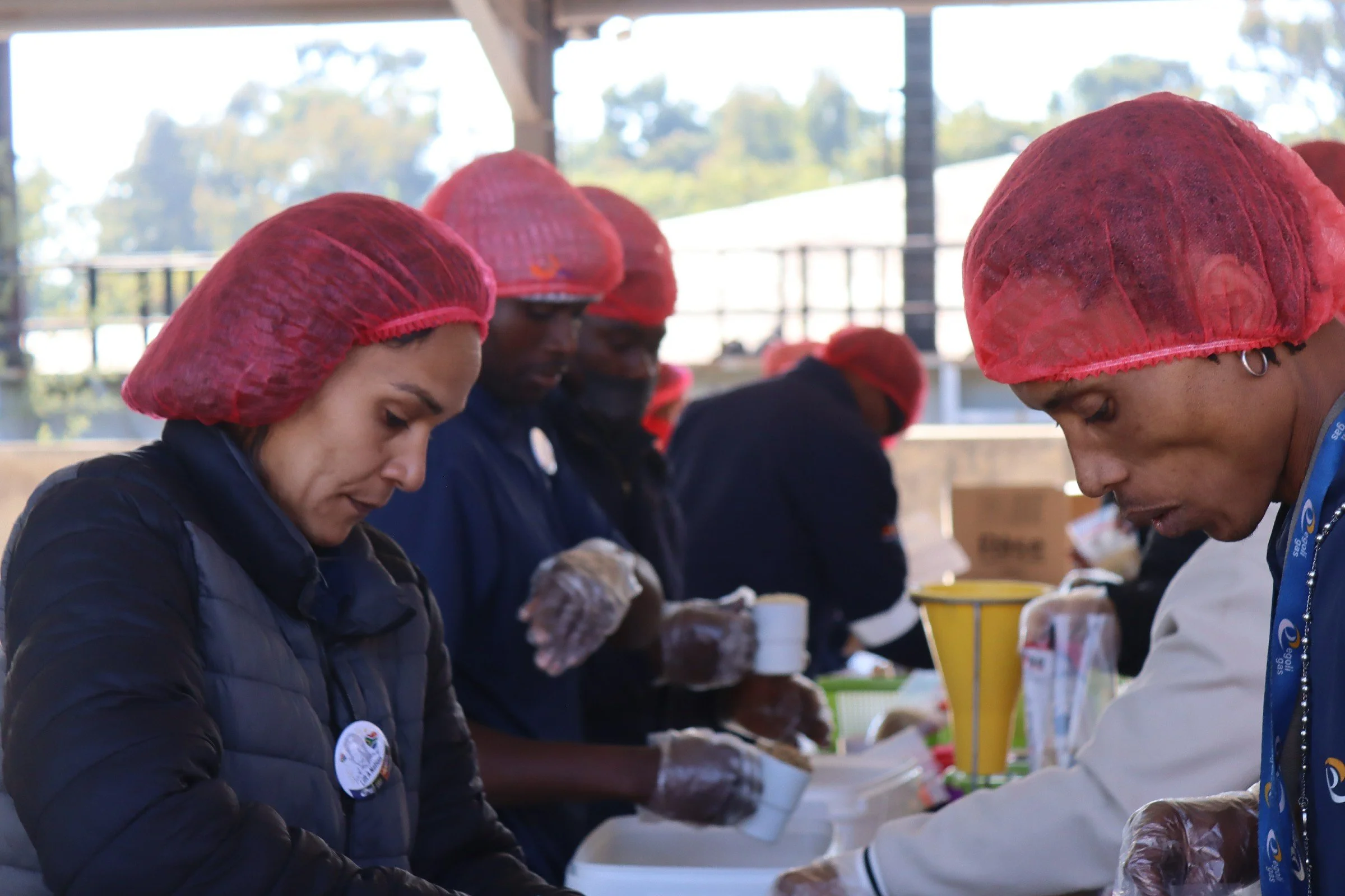 People working together, wearing red hairnets, in a communal setting, possibly preparing food or participating in a group activity.