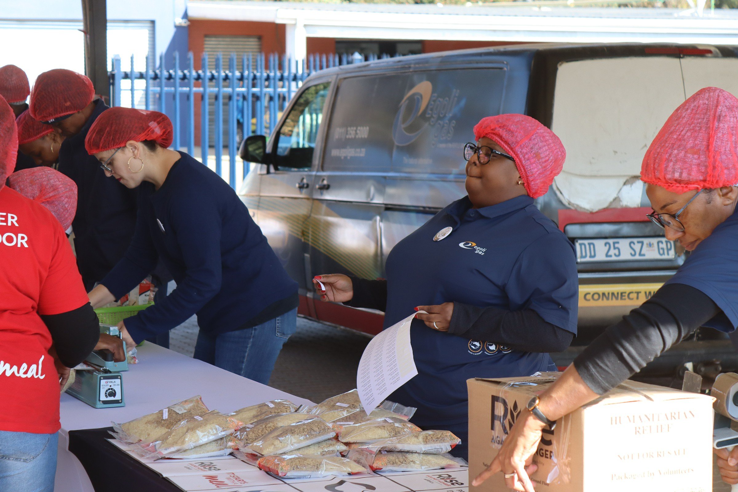 People participating in a food distribution event, wearing red hairnets, gathered around a table with packaged food, with a vehicle and blue fence in the background.