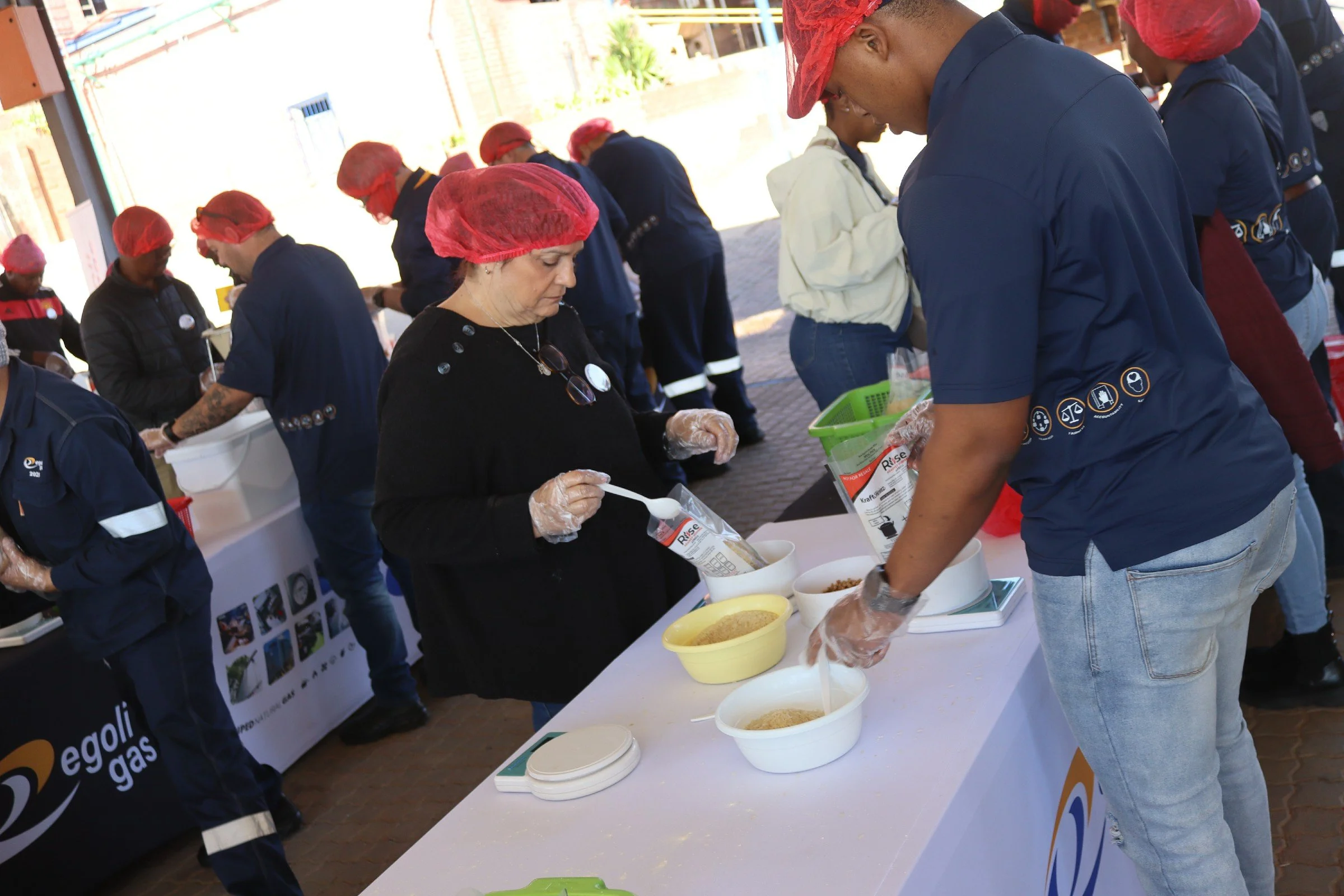 People standing around a table filling bags with grains or food at an outdoor event, some wearing red hairnets and gloves.