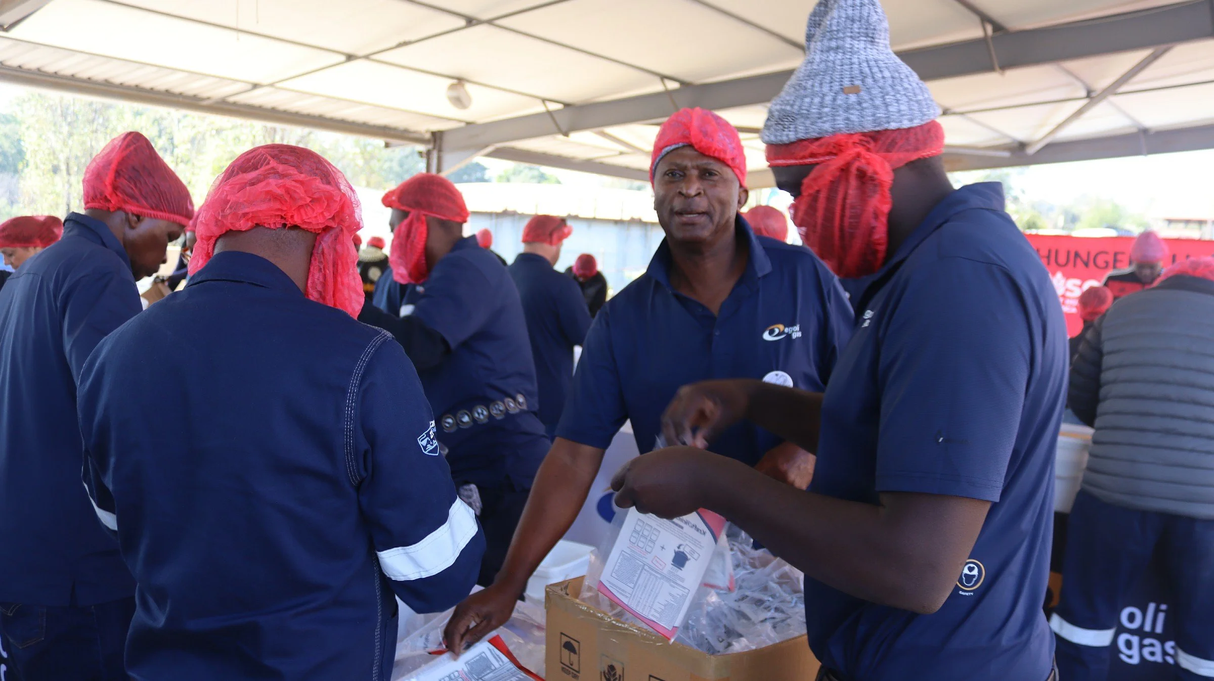 People wearing blue uniforms and red hairnets, gathered under a tent, packing items and talking, possibly at a community event or food distribution.
