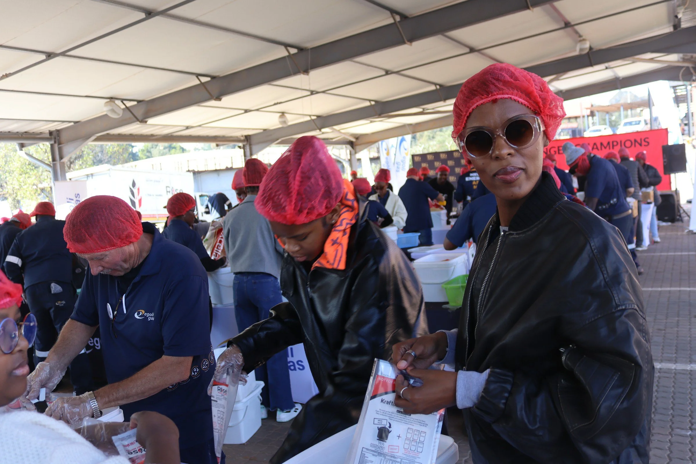 People working under a large tent, wearing red hair nets, in an organized setting.