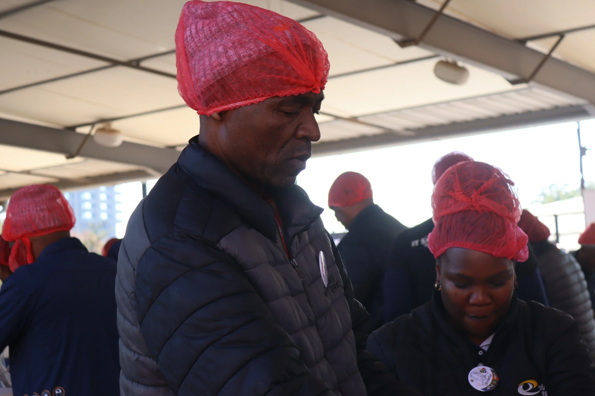 A group of people wearing red hairnets and dark jackets, standing with heads bowed in an indoor setting with a metal ceiling and bright natural light.