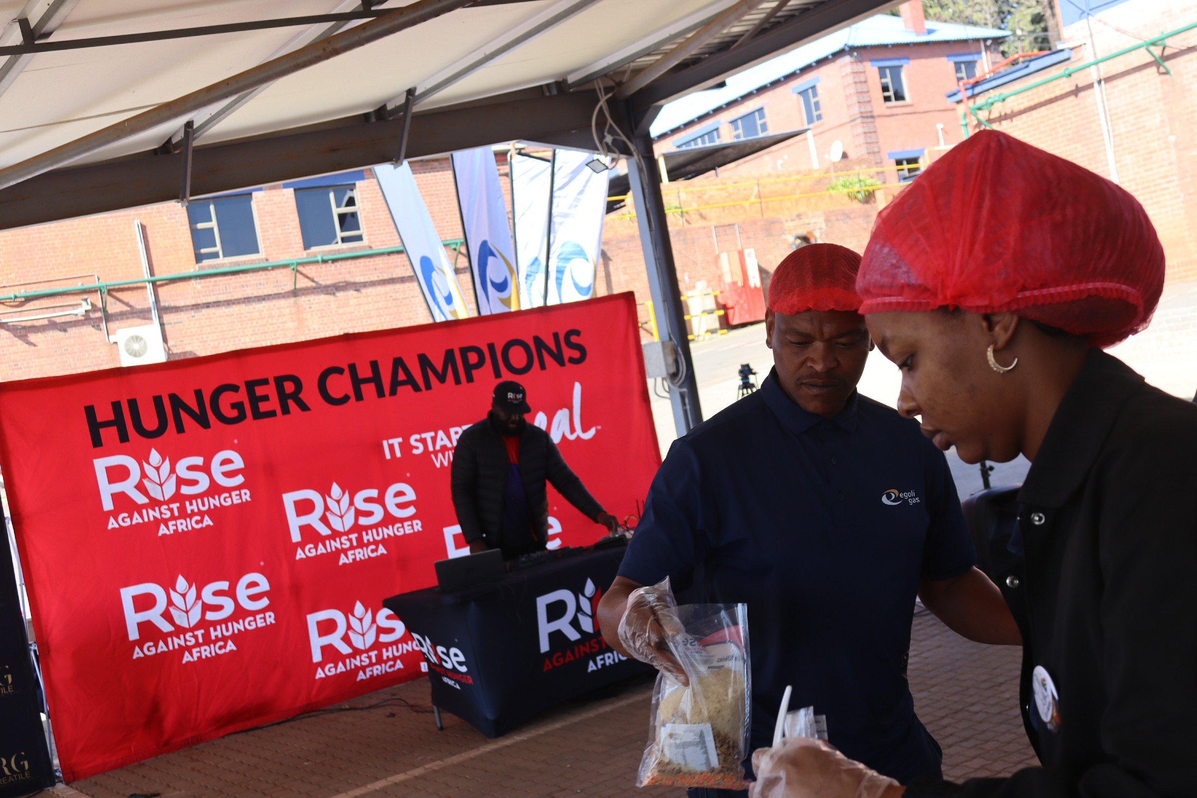People at a food distribution or charitable event for raising awareness against hunger in Africa, with a backdrop of blue and white banners and a red banner stating "Rise Against Hunger Africa".