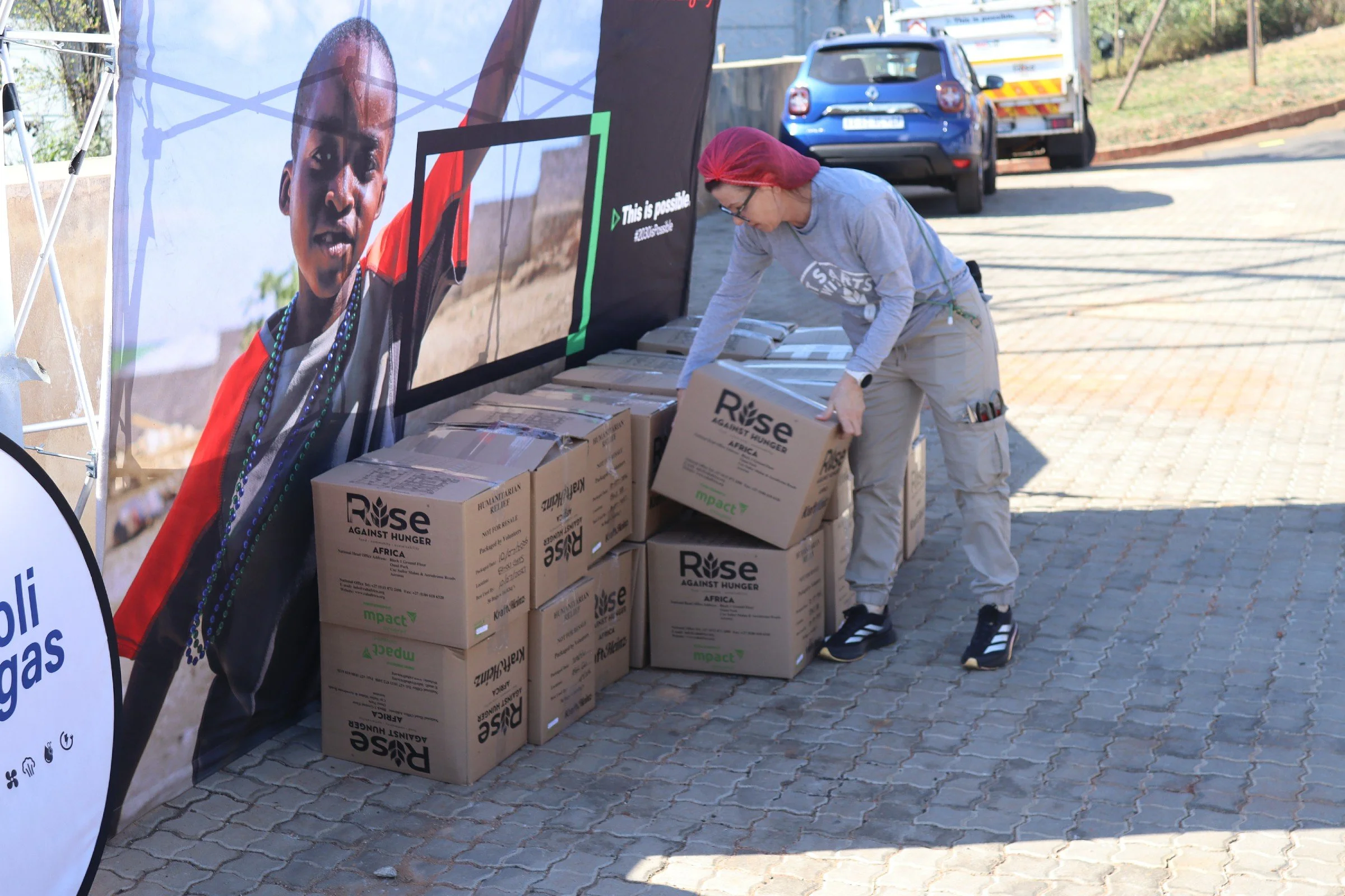 A woman with a red headscarf and gray sweatshirt loading boxes onto a curb next to a large rally banner for Rise Against Hunger, featuring a child's face and the slogan "This is possible".