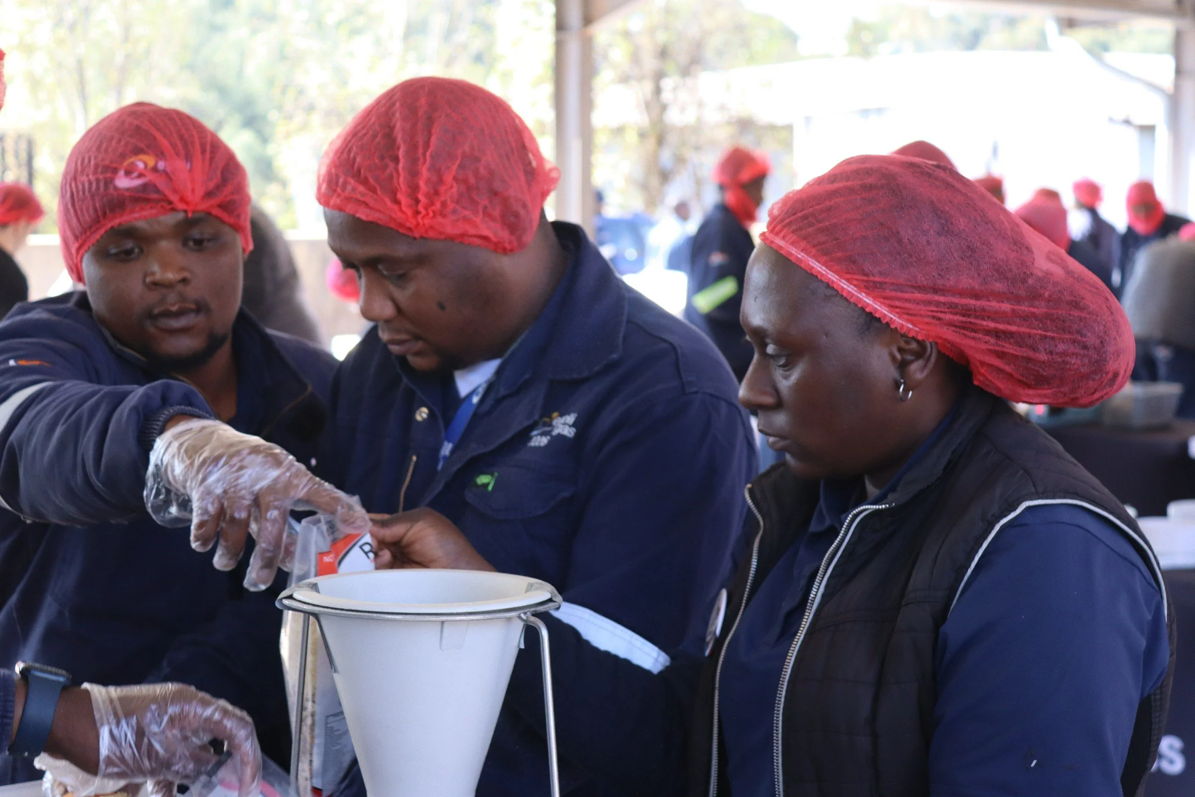 Several people wearing red hairnets and dark clothing gather around a white container, working together at an outdoor event with other people in the background.