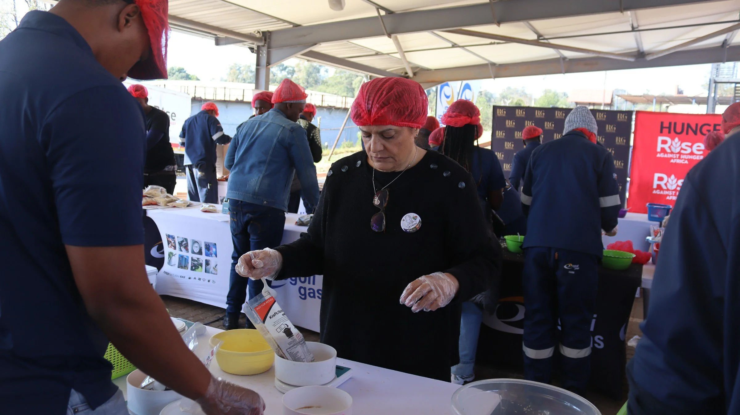 People wearing red hairnets and gloves preparing and serving food at an outdoor event, with a banner that reads 'Hunger Rise Against Hunger Africa' in the background.