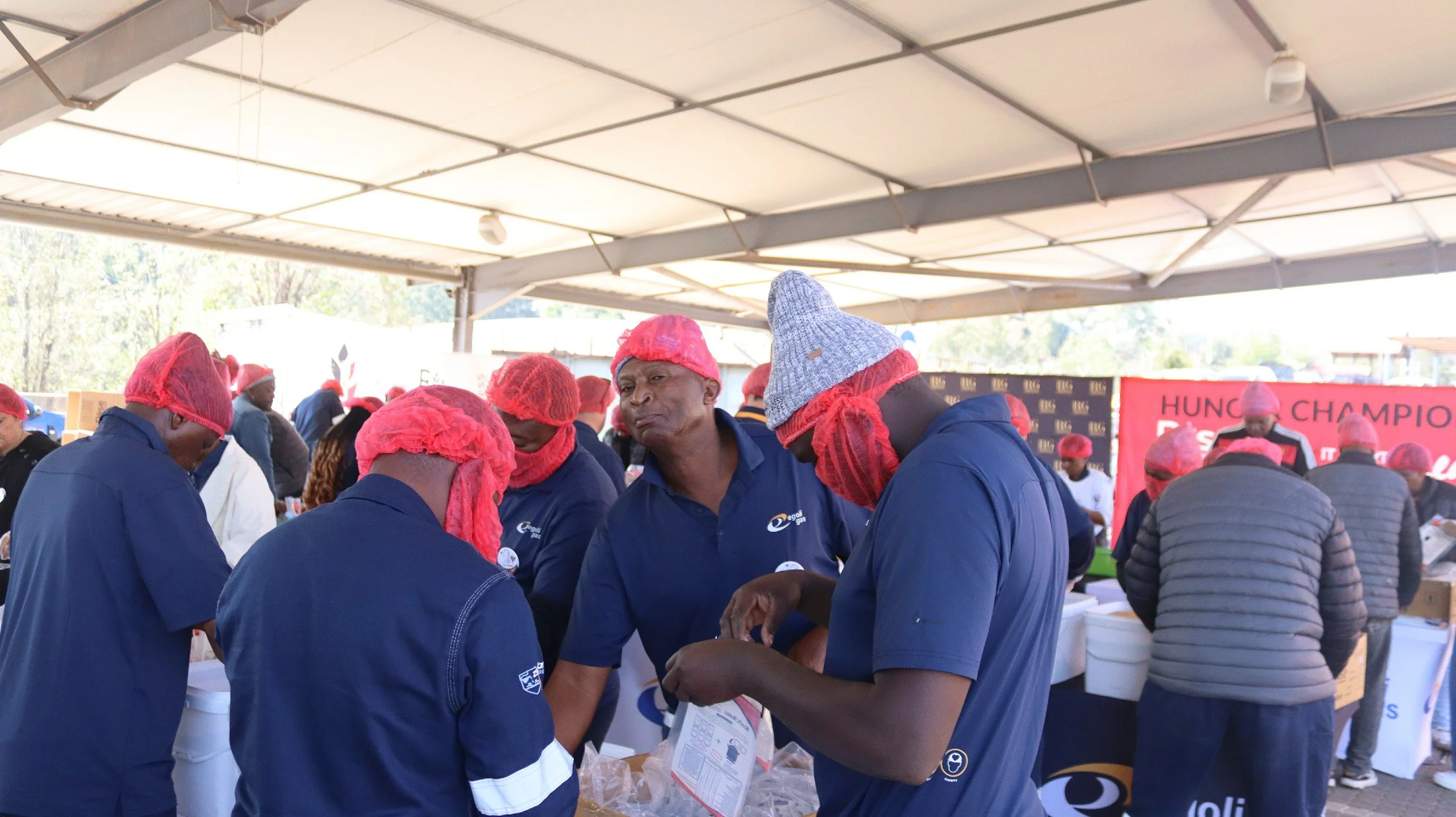 Group of people wearing blue shirts and red hairnets under a large canopy, engaged in a collaborative activity at an outdoor event.
