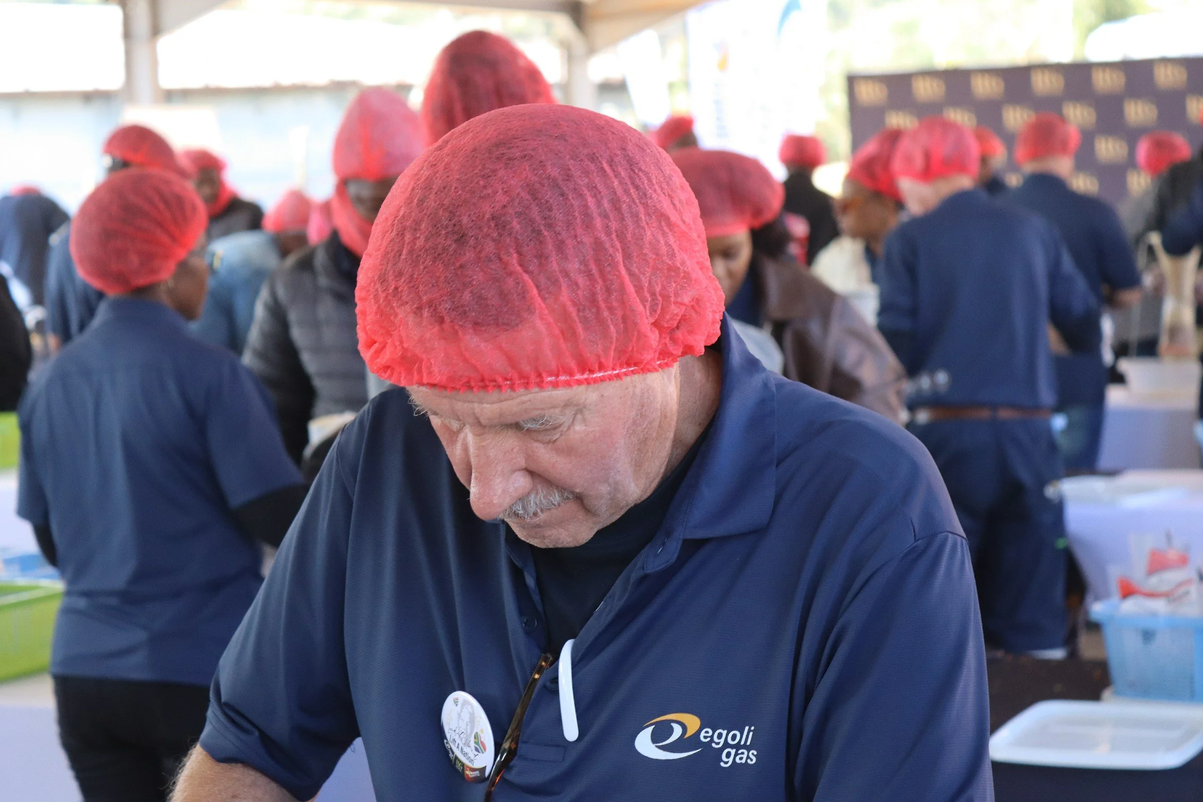 A man wearing a red hair net and a blue shirt with the logo 'Egoli Gas' is participating in an event with several other people, all wearing red hair nets. They appear to be working together at a table or station under a covered outdoor area.