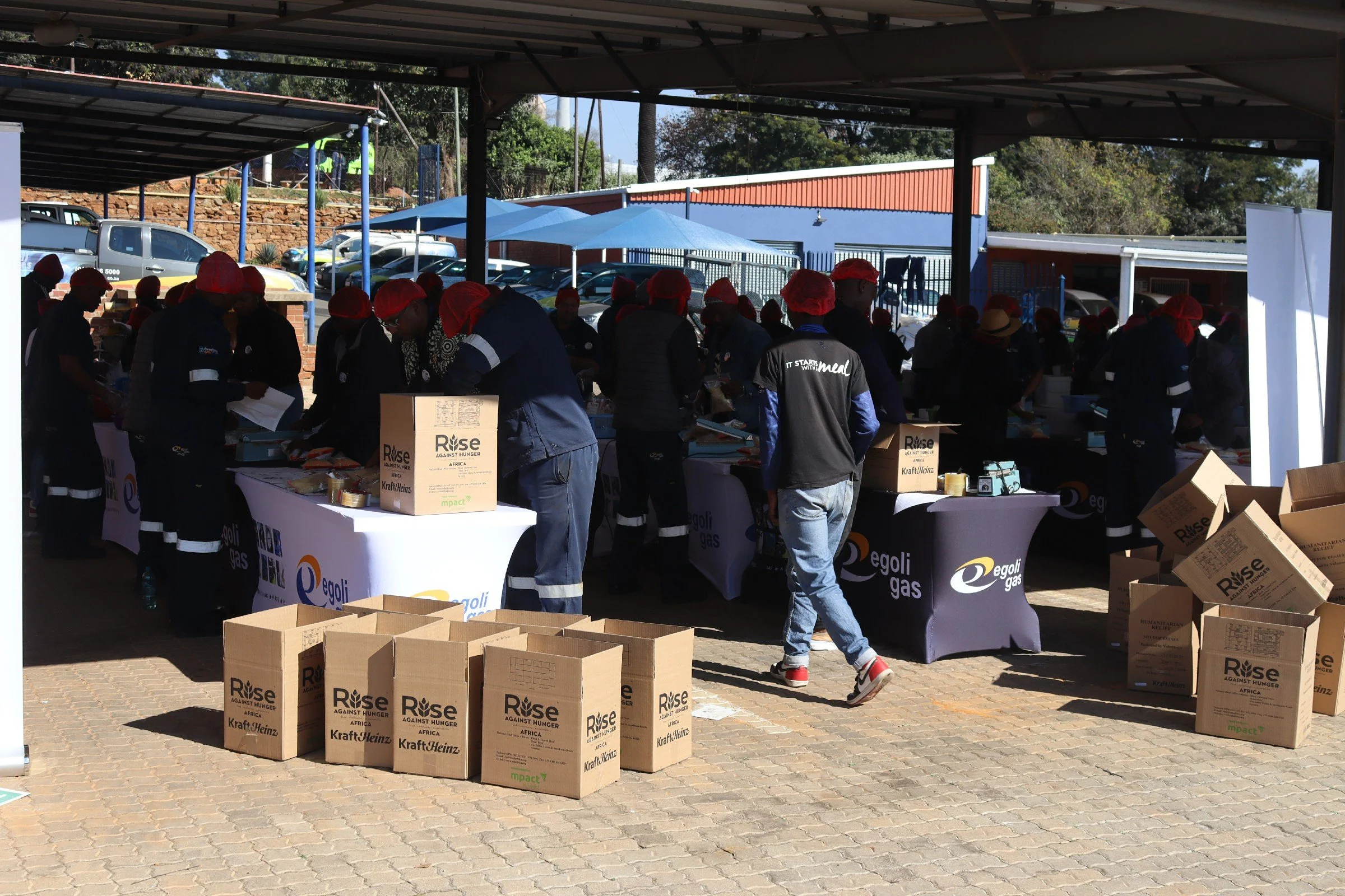 Several people wearing red hairnets are working at tables under a shaded outdoor structure, organizing food donations with cardboard boxes labeled 'Rise Against Hunger Africa' situated around the area.