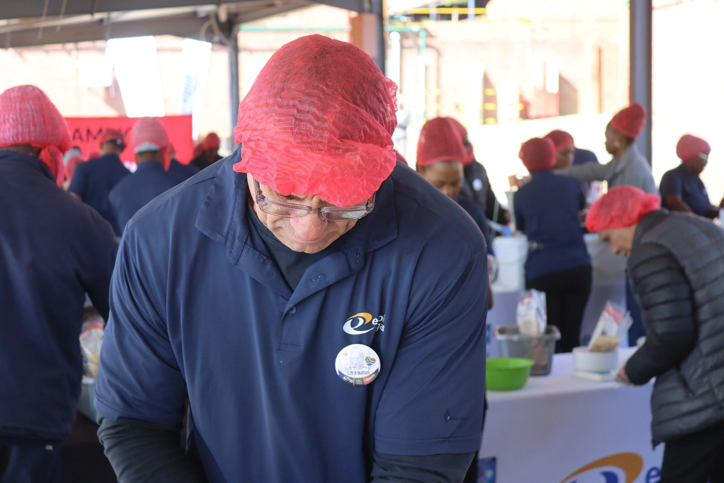 Several people wearing red hair nets and navy blue uniforms working at a table in an industrial setting, with a woman in the foreground wearing glasses and a badge reading "Lift a Nation."