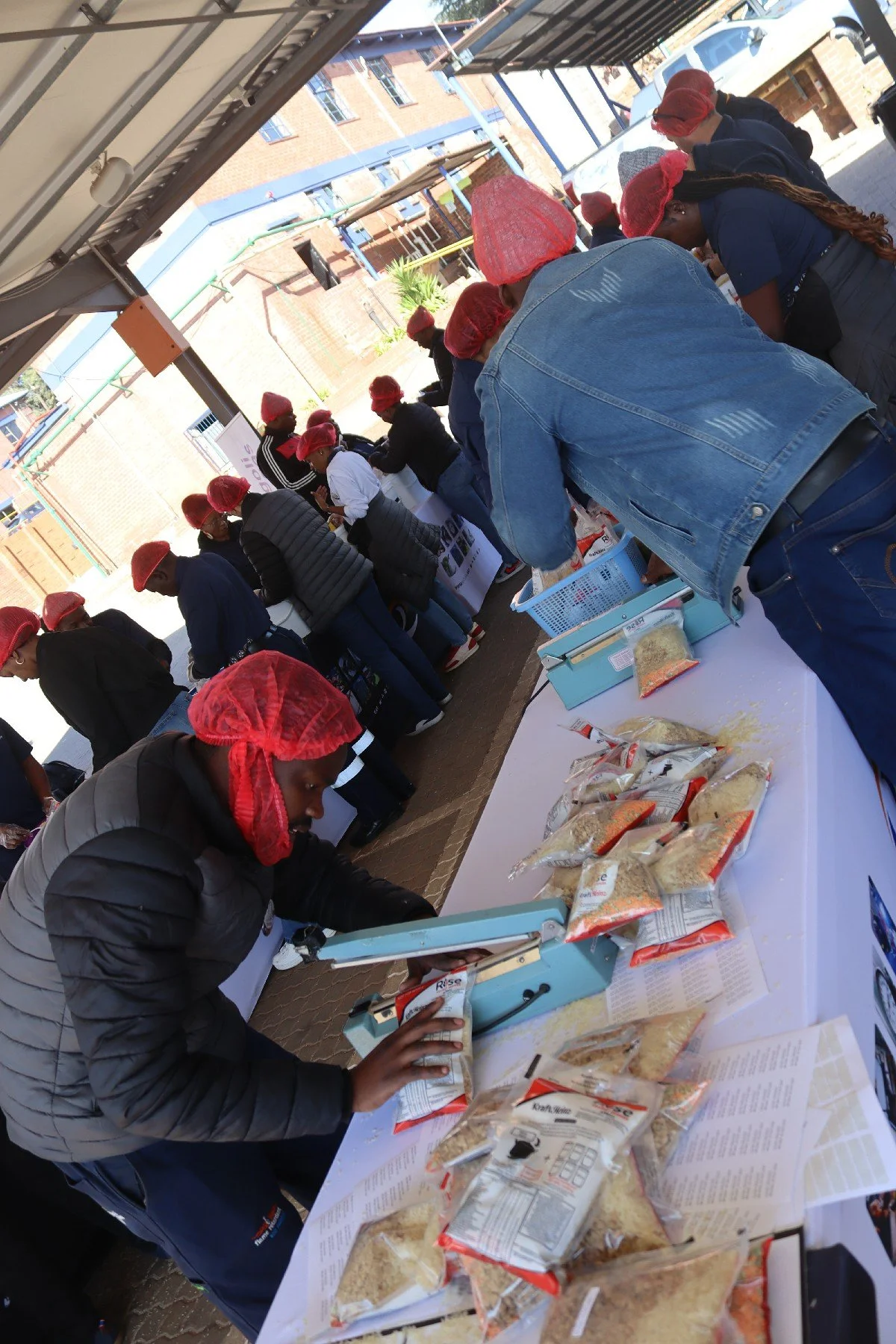 People gathered around a table at an outdoor event, packaging grains or rice into bags. Some individuals are wearing red hair covers or bandanas, and the setting appears to be a community or volunteer activity.