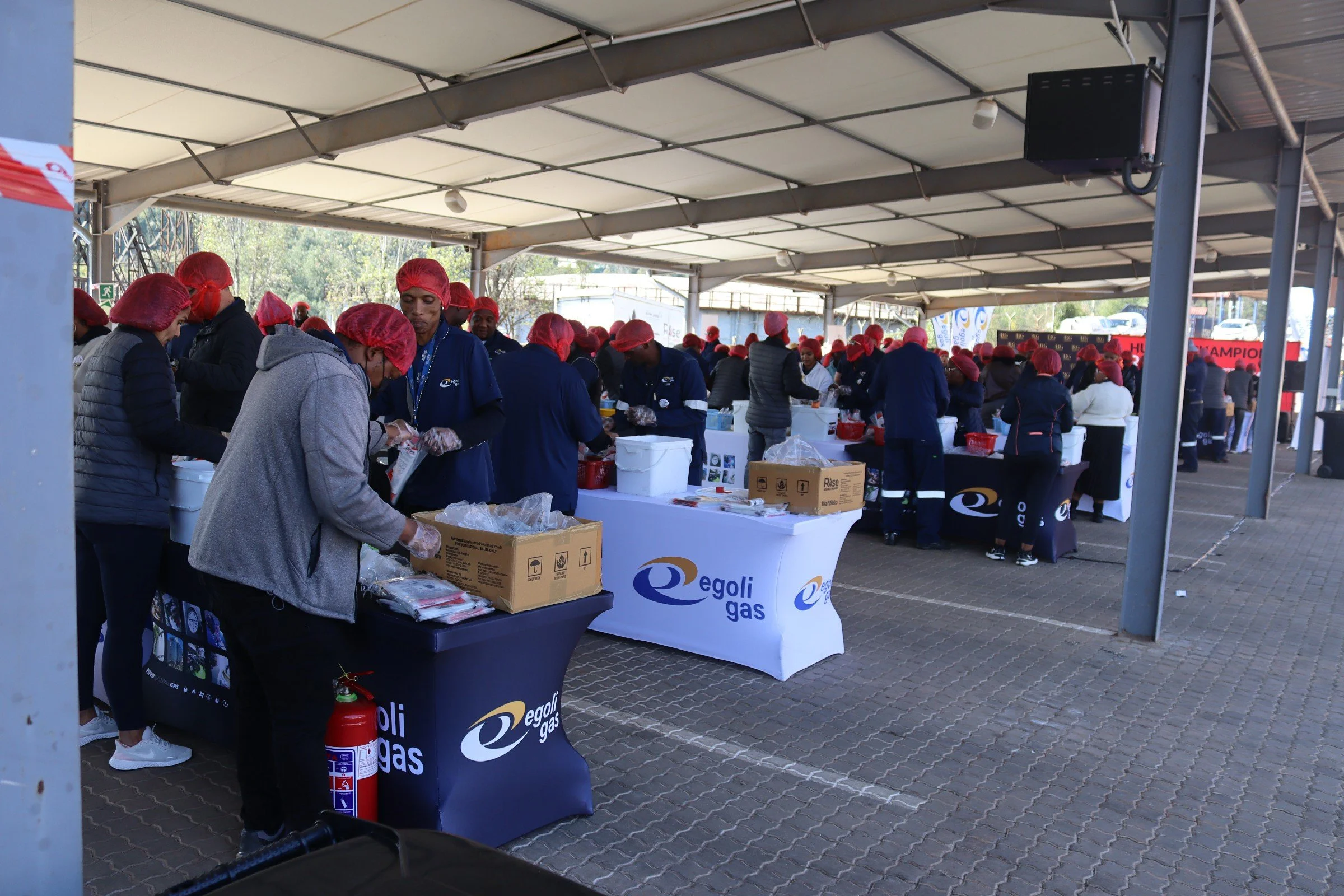 People working at a food service event, wearing red hair nets under a large outdoor canopy, with tables displaying Egoli Gas branding and boxes of supplies.